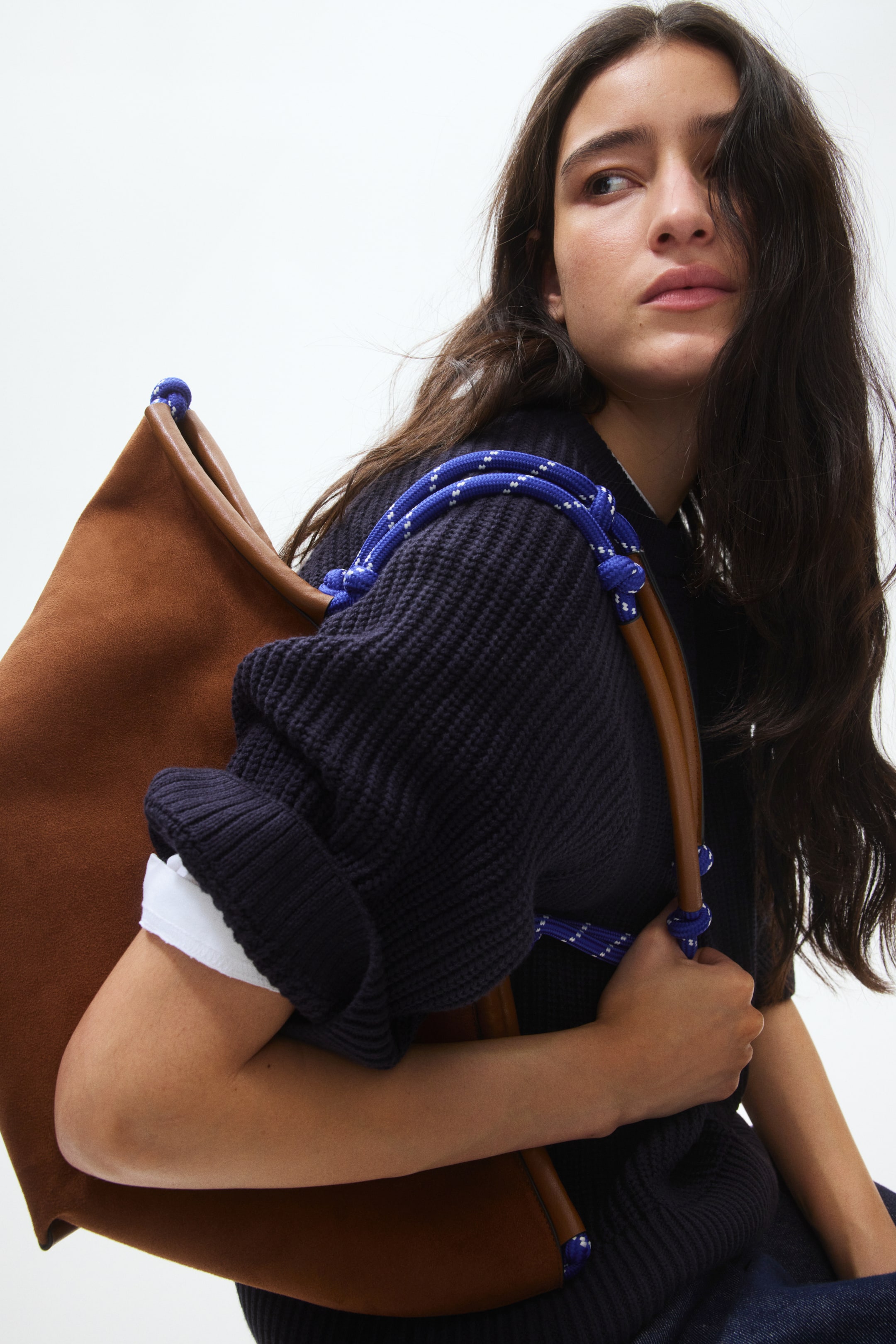 View larger image: A brown suede bag featuring smooth brown leather trim along its top edge, with blue and white striped rope straps tied around the trim and resting on a dark knit jumper.