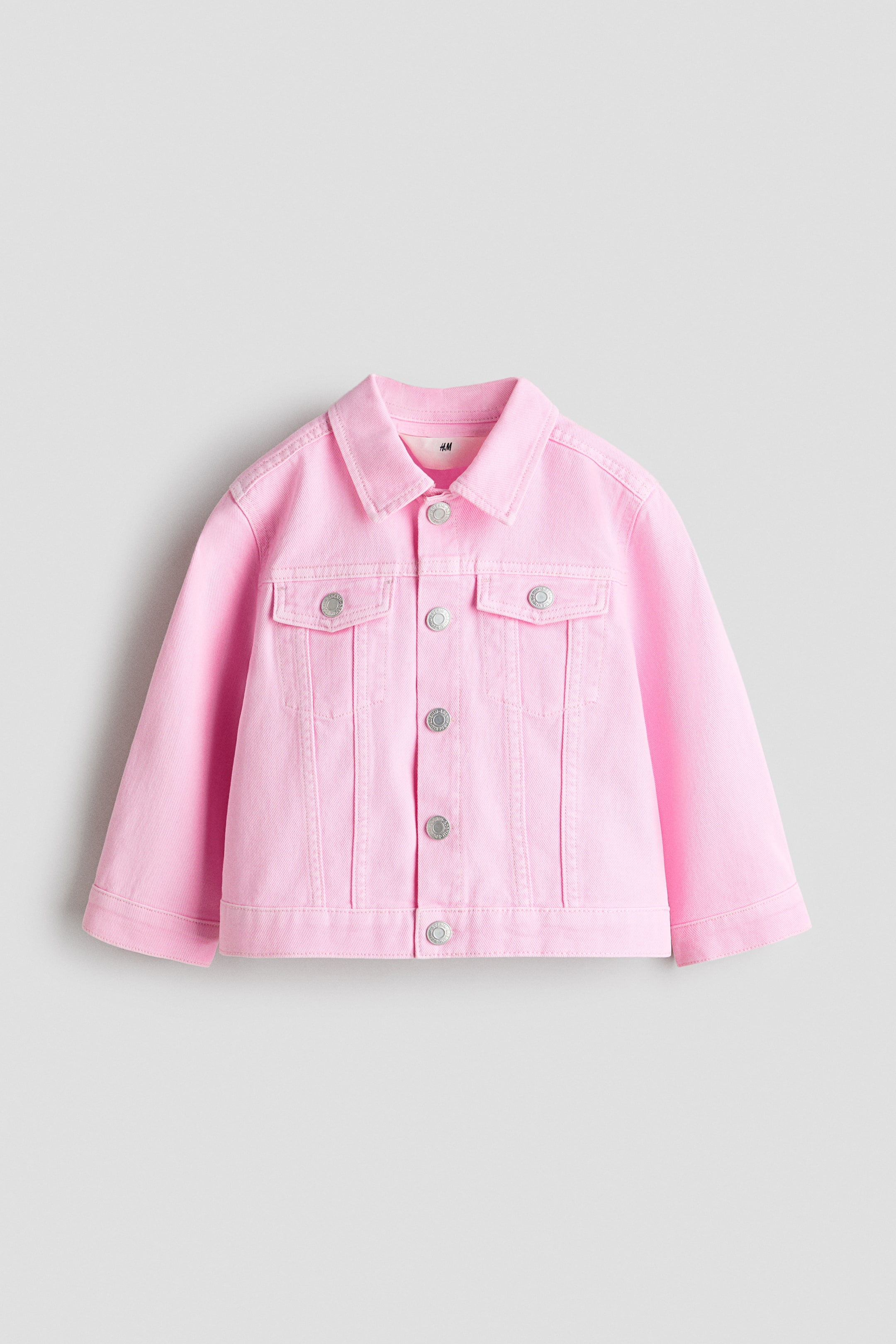 Visa större bild: En klassisk jacka i rosa mjuk bomullsdenim visas framifrån, med en nedvikt krage, tryckknappar längs fram och tryckknappar på ärmsluten. Dekorativa bröstfickor syns också, var och en med en klaff med tryckknapp.