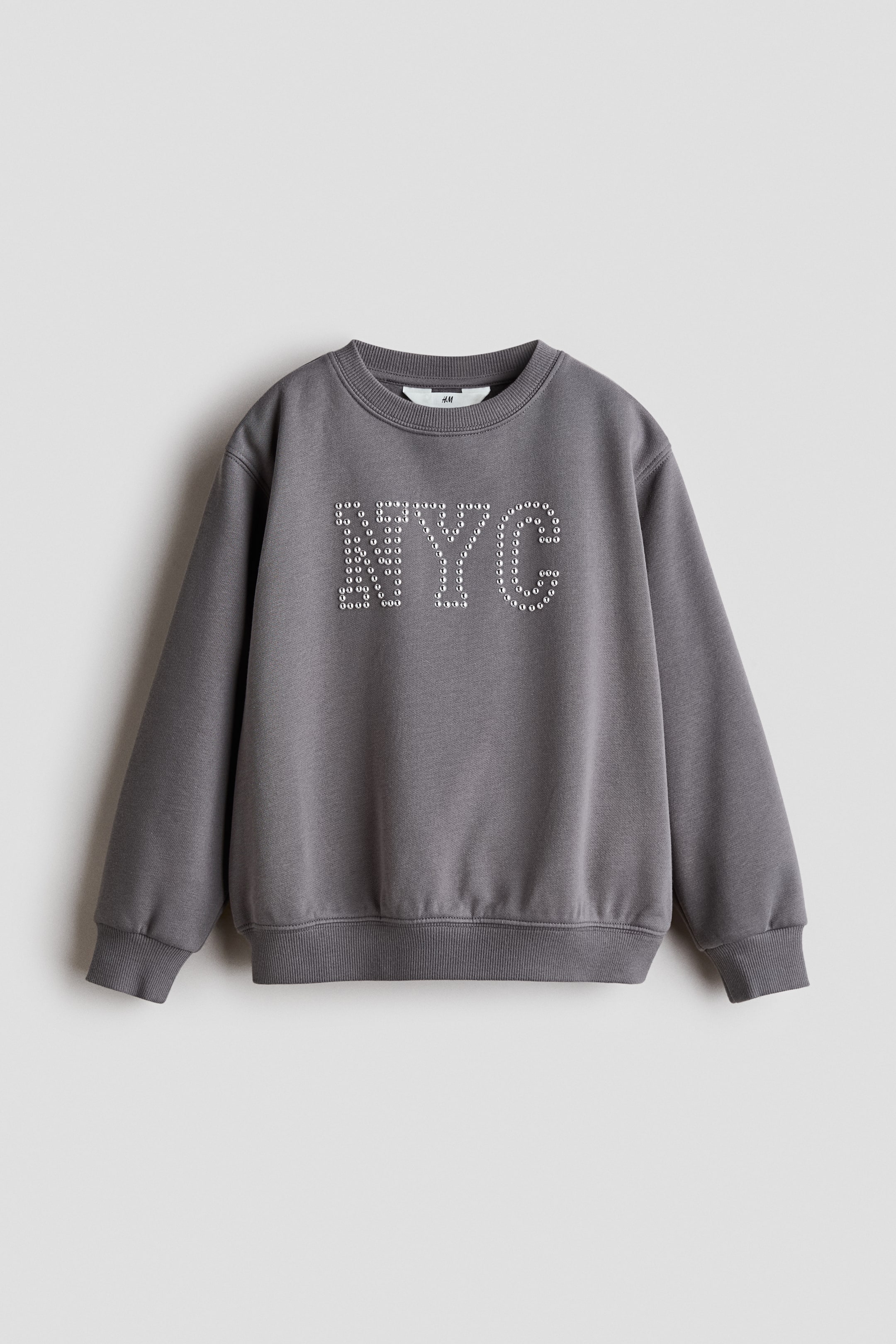 View larger image: A grey sweatshirt is displaying a front-facing view, featuring a crew neck, ribbed cuffs, and a ribbed hem. The front of the sweatshirt showcases "NYC" in large capital letters, formed by numerous small, round, silver-coloured studs.