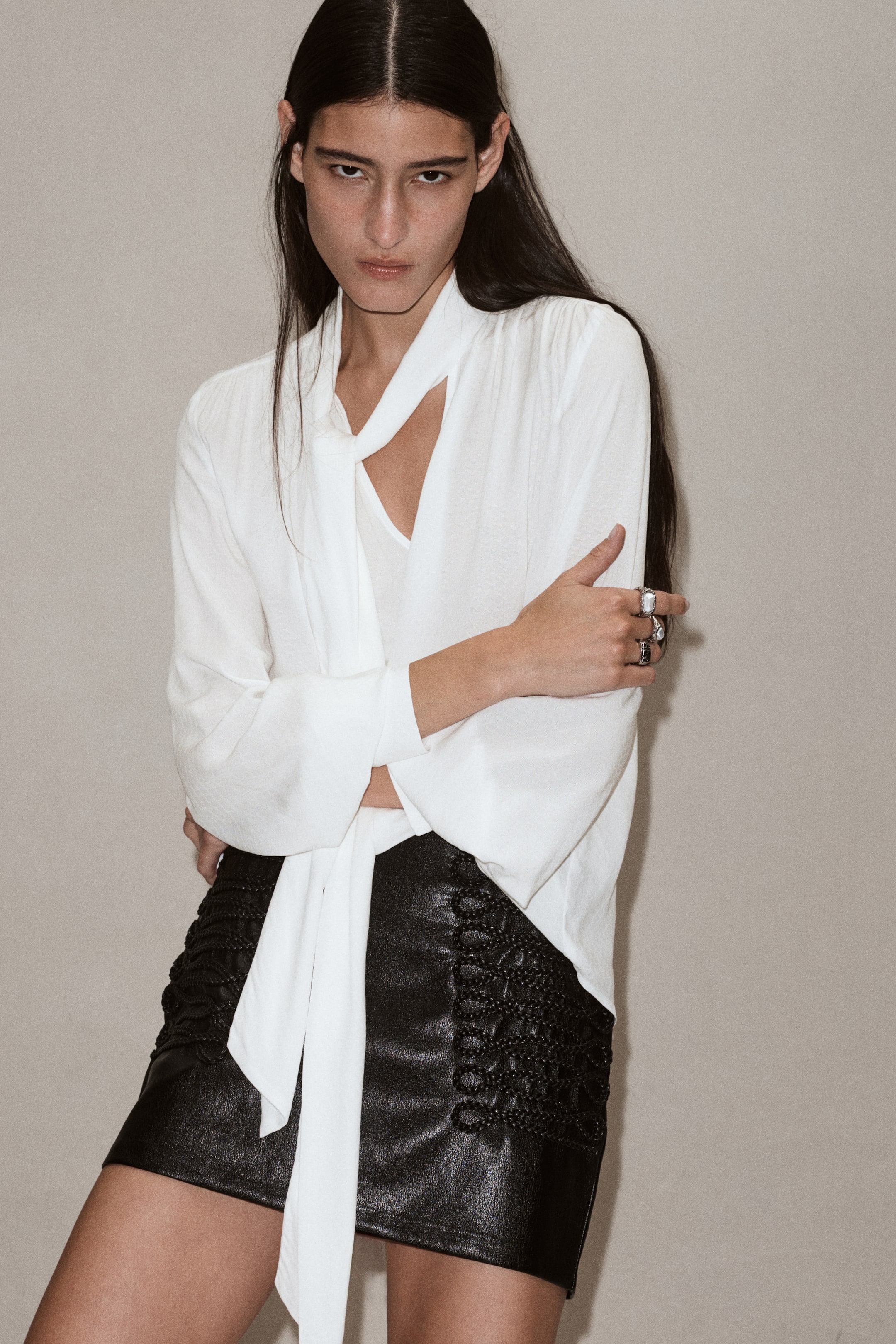 View larger image: A white, loose-fit blouse in woven fabric is being worn by the model, featuring a draped scarf collar, a V-neck front opening, and long balloon sleeves. The blouse is styled with a textured black mini skirt, while the model poses with folded arms.