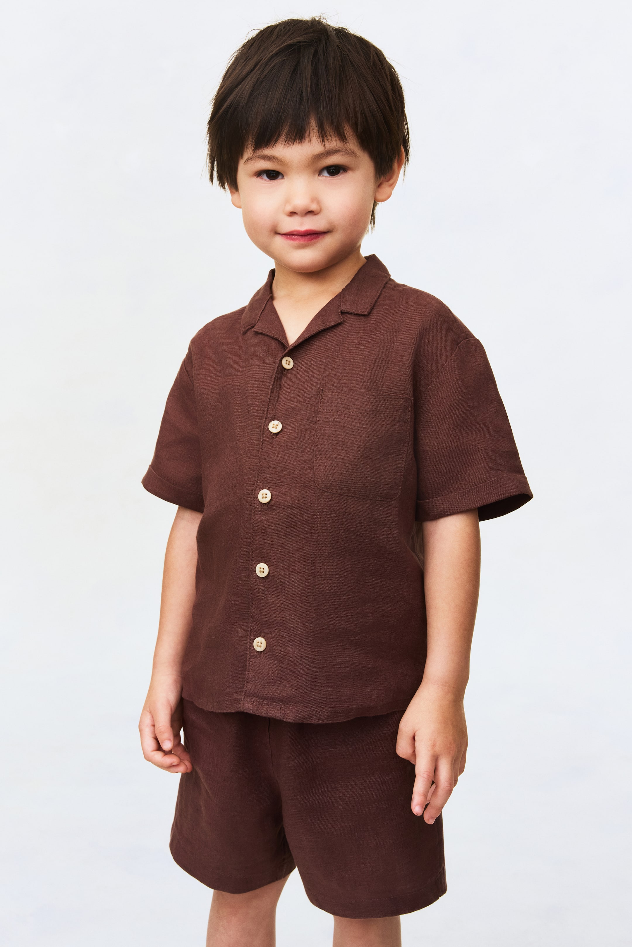 View larger image: A dark brown short-sleeved shirt and matching shorts set is displayed facing forward. The shirt features a camp collar, a full button placket with light-coloured buttons and a single chest pocket, while the coordinating shorts have a relaxed fit.