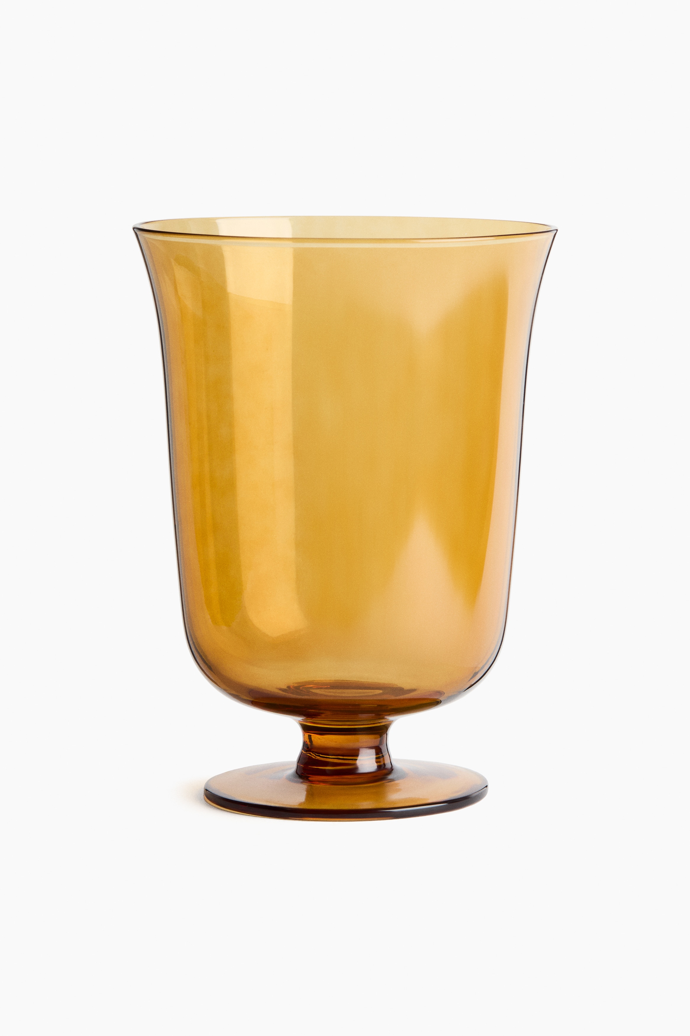 A glass footed vase made of translucent amber glass is shown from the front, featuring a gently flared opening, a rounded body, and a short stem on a flat circular base. Several leafy branches with reddish-purple berries are resting inside the water-filled vase, highlighting its transparency and timeless design.