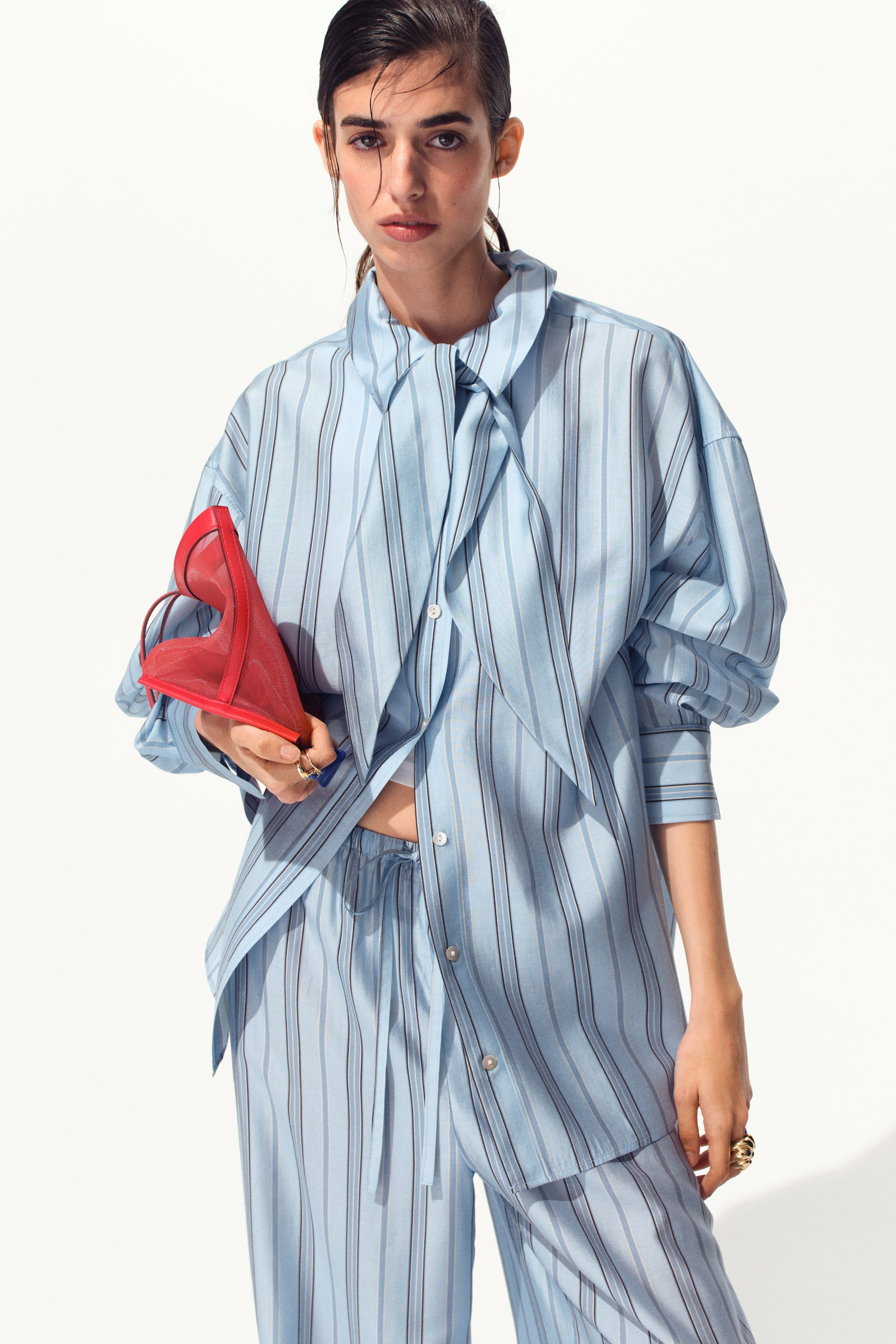 View larger image: A light blue striped pajama-style shirt and matching pants are showcased, viewed from the front. The oversized shirt displays varying blue and dark vertical stripes, a pussy-bow collar, and buttoned cuffs on its long voluminous sleeves, and it is worn partially unbuttoned over a white undergarment. The matching pants feature a drawstring waist and a relaxed fit.