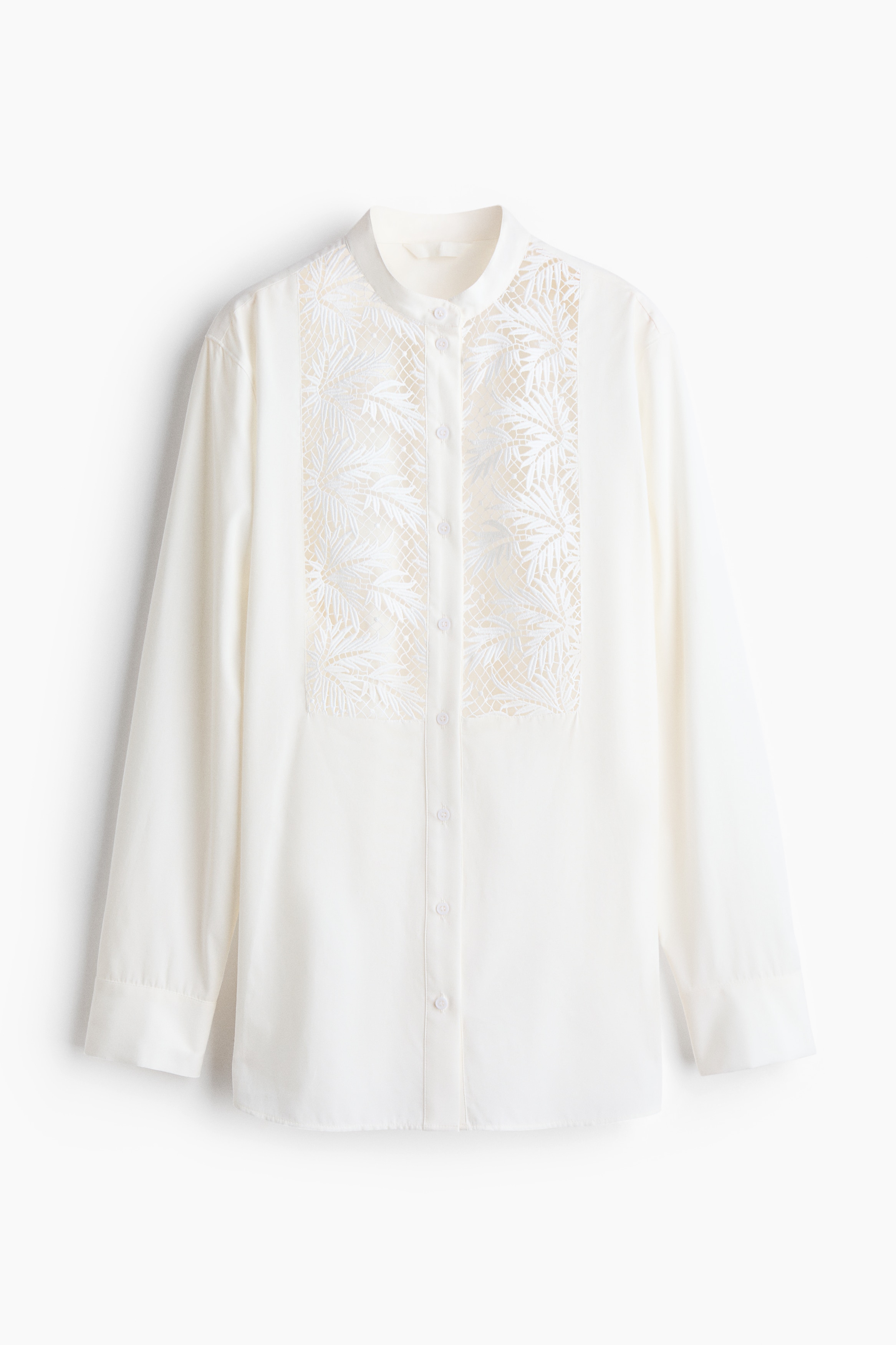 En oversized skjorte i en lys vevstruktur bæres av modellen, med bandkrage og knappelukking foran. Skjorten er detaljert med et stort, intrikat brodert blondestykke foran, med et bladlignende åpent mønster.