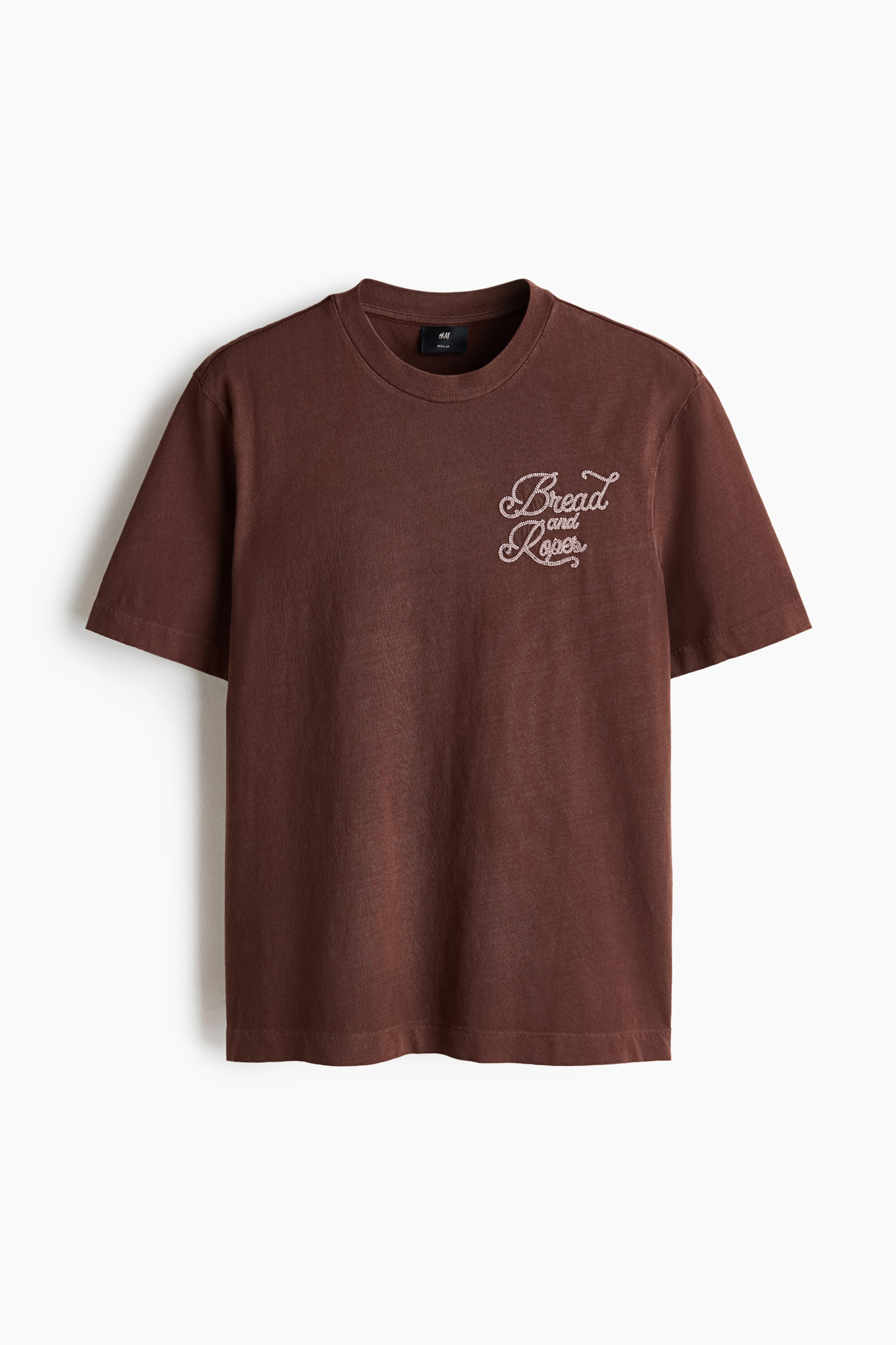 View larger image: A brown t-shirt is facing front, featuring a washed brown fabric, a crew neck, and short sleeves. The phrase "Bread and Ropes" is decorating the left chest in a light-coloured, embellished script font.