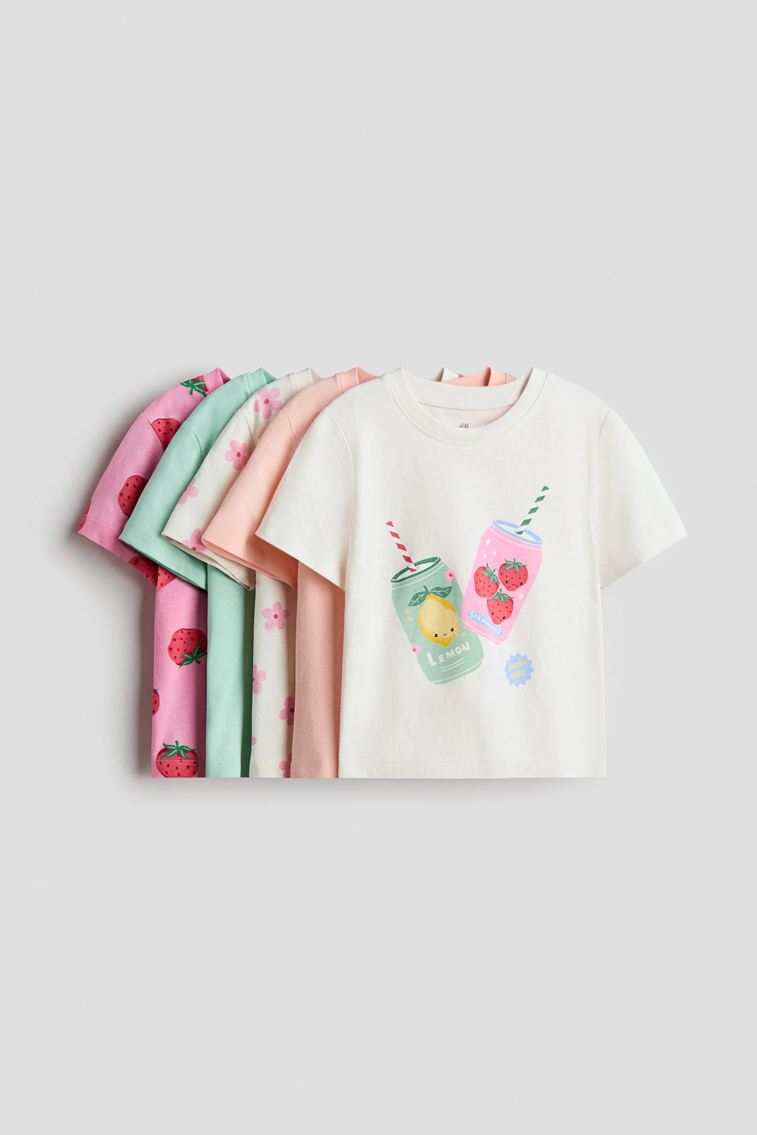 Vijf T-shirts zijn gestapeld, waarbij het voorste off-white T-shirt naar voren is gericht en een grafische print van twee cartoon drankblikjes toont. Alle T-shirts hebben korte mouwen en geribbelde boorden rond hun halslijnen, terwijl de gedeeltelijk zichtbare T-shirts erachter patronen vertonen van roze aardbeien, mintgroen, crème met roze bloemen en effen perzik.
