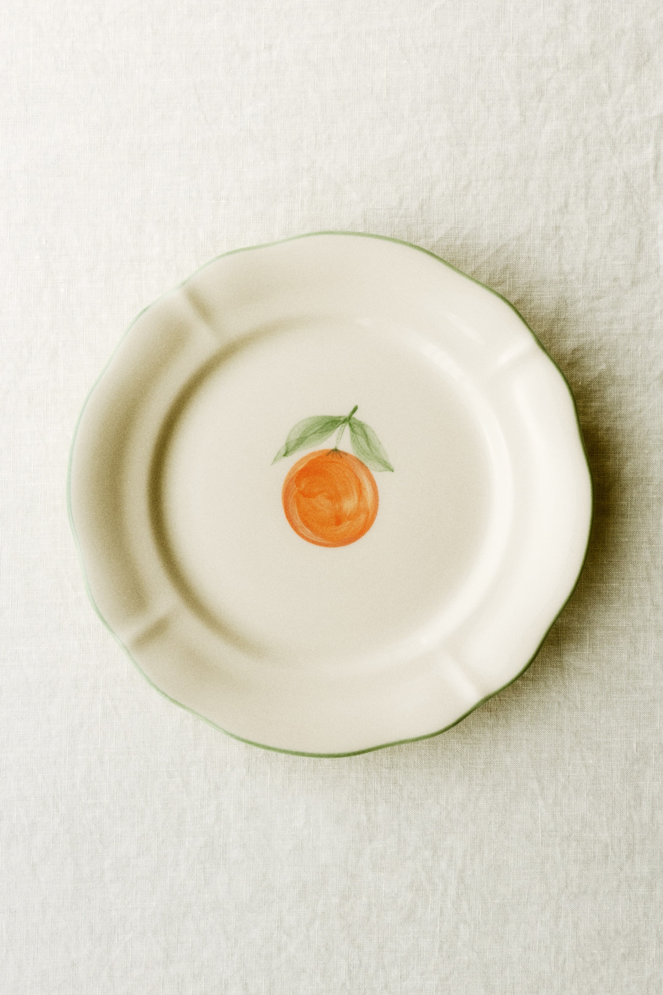 Zobacz większy obraz: Biały ceramiczny talerz z falistym brzegiem i zieloną obwódką, z ręcznie malowaną pomarańczą z zielonymi liśćmi w centrum, widziany z lekko podwyższonej perspektywy.
