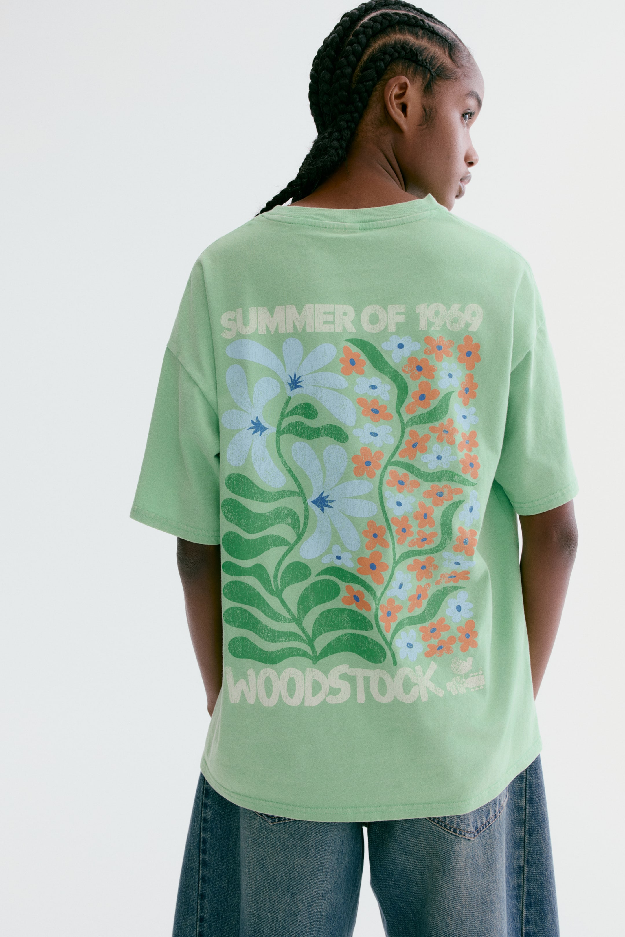 Agrandir l'image: Le t-shirt vert clair est présenté de dos, arborant une coupe ample et des manches courtes. Un grand imprimé graphique vieilli recouvre le dos, affichant "ÉTÉ DE 1969" en haut, "WOODSTOCK" en bas, et un motif floral central avec des éléments bleus, orange et verts.