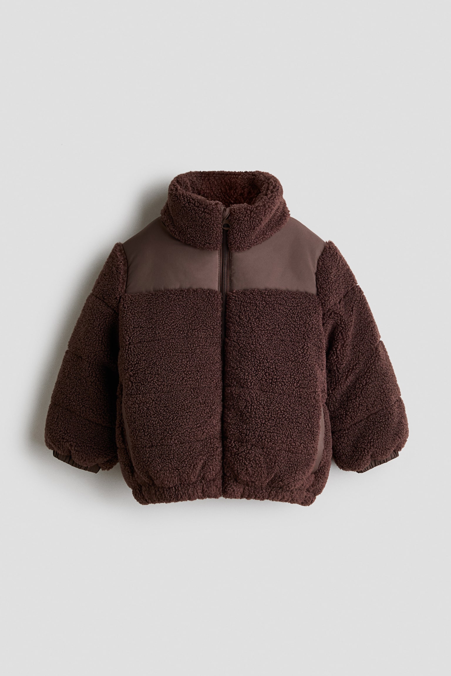 Una chaqueta puffer sherpa marrÃ³n oscuro para niÃ±o/a aparece de frente, mostrando un cuerpo y unas mangas de forro polar texturizado contrastados con paneles lisos de tejido en la parte superior del pecho. La chaqueta presenta un cuello alto tipo chimenea, cierre frontal de cremallera completa y puÃ±os y bajo elÃ¡sticos.