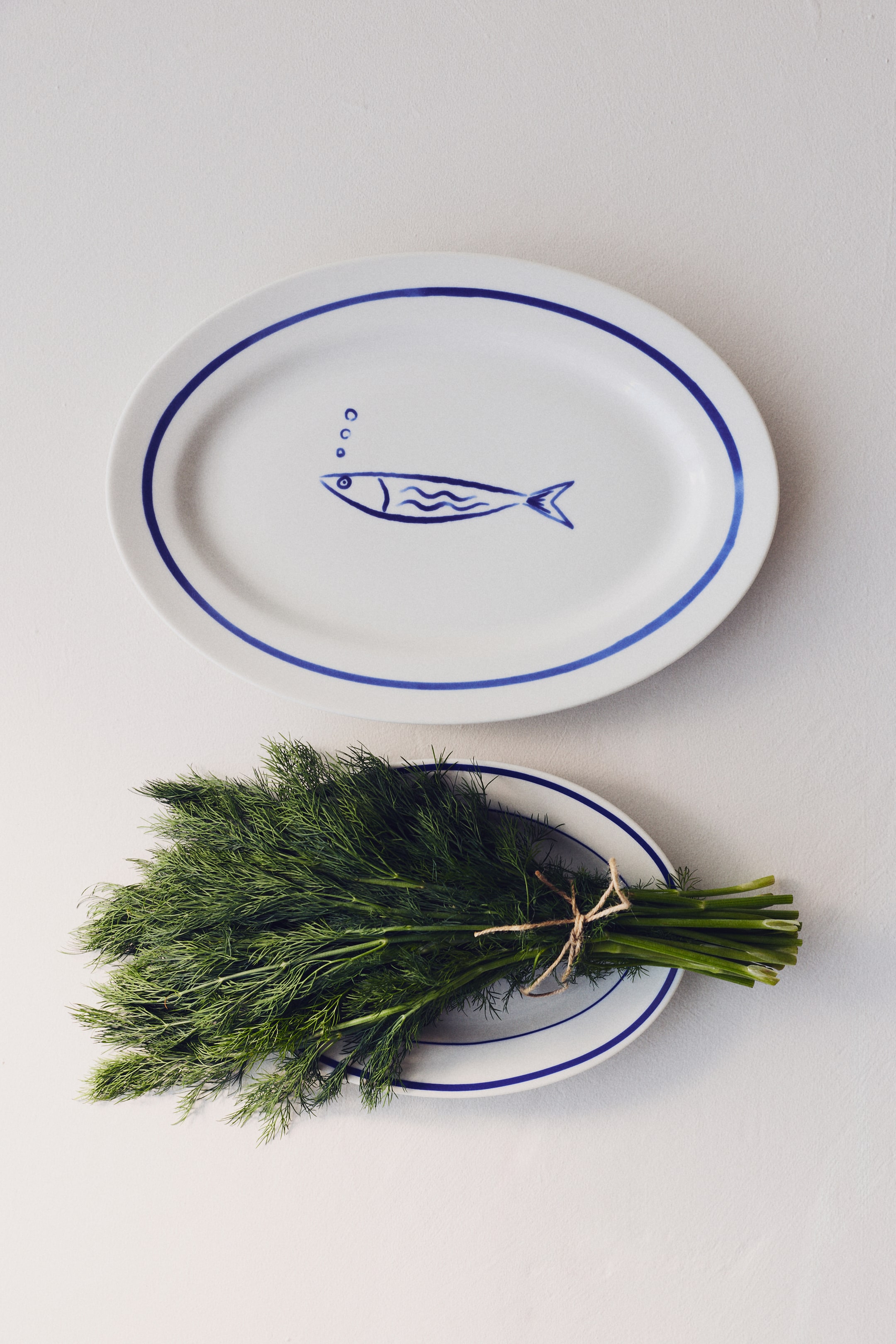 View larger image: An oval white serving platter is lying flat, featuring a blue rim and a central hand-drawn blue fish design. The fish is facing left, with three small blue circles representing bubbles above its mouth.