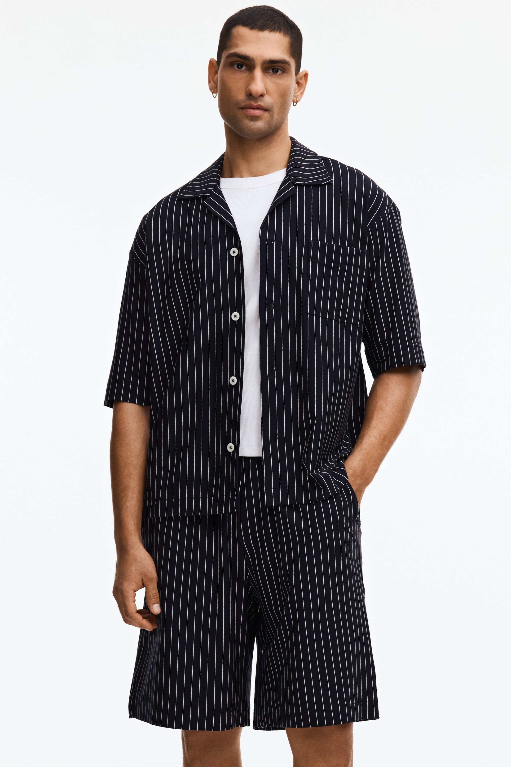 Ingrandisci l'immagine: Una camicia blu scuro a maniche corte con sottili righe bianche è indossata aperta sopra una t-shirt bianca, caratterizzata da un colletto a revers e una tasca applicata sul petto. Sono indossati anche pantaloncini blu scuro abbinati con sottili righe bianche, formando un set coordinato.