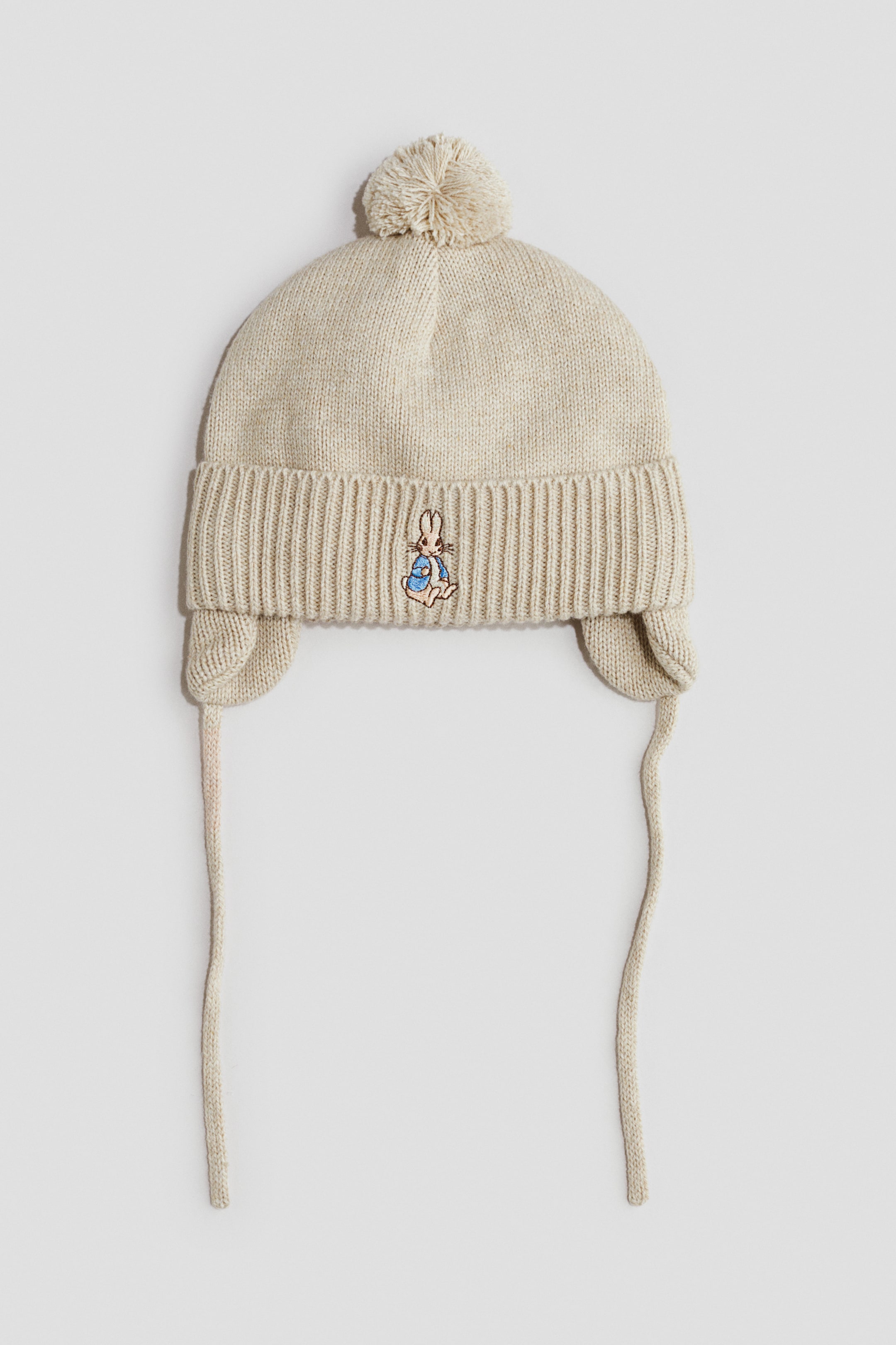 View larger image: A light beige knitted hat features a pom-pom on the crown and a wide ribbed band. It has ear flaps with knitted ties extending downwards, and a small embroidered rabbit character adorns the front of the ribbed band.