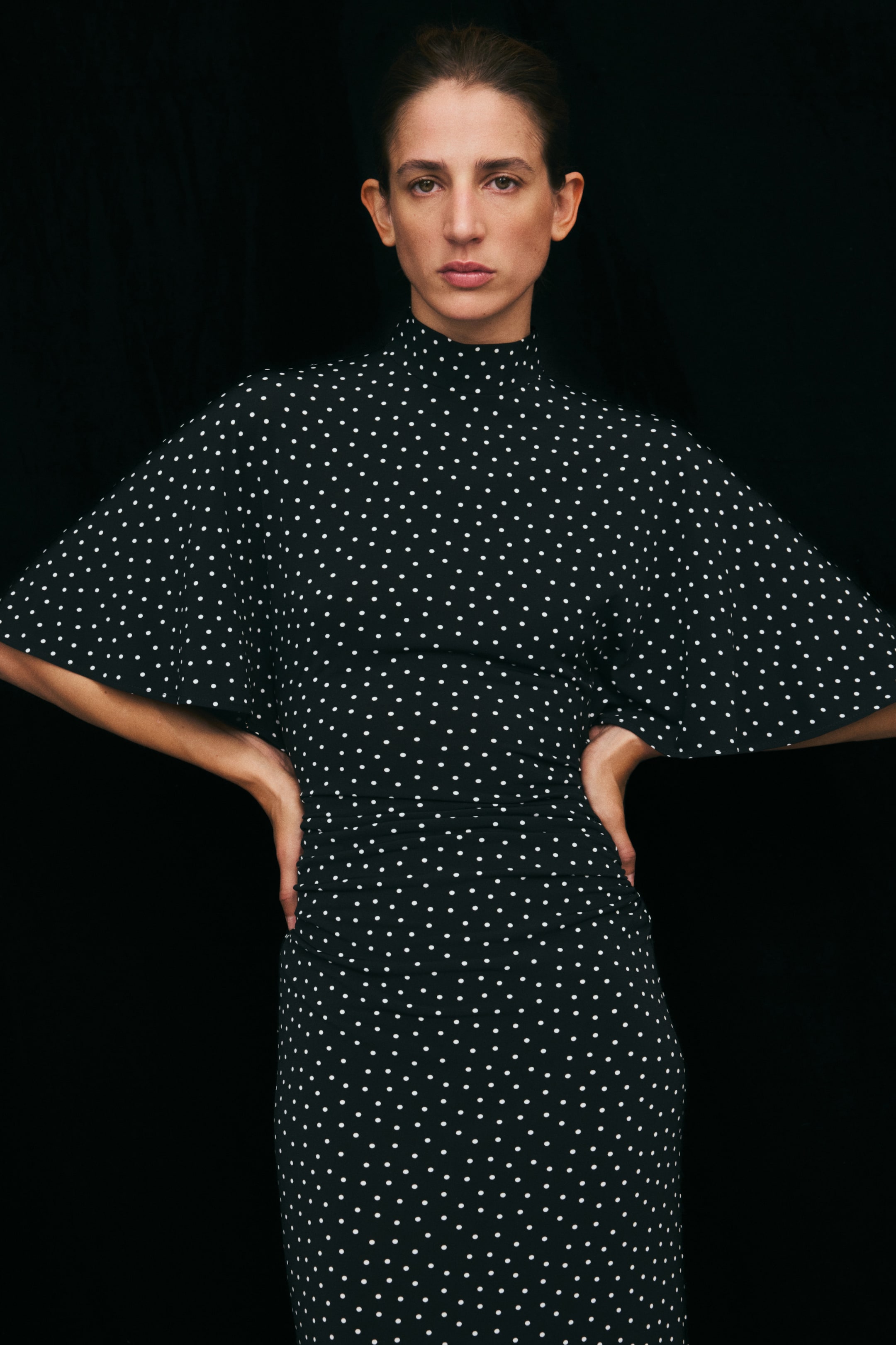 Ampliar la imagen: La modelo lleva un vestido negro de jersey con lunares blancos, que presenta cuello alto, mangas amplias tipo mariposa a la altura del codo y cintura fruncida. La modelo posa con las manos apoyadas en las caderas, destacando la cintura definida del vestido y las mangas con caída.