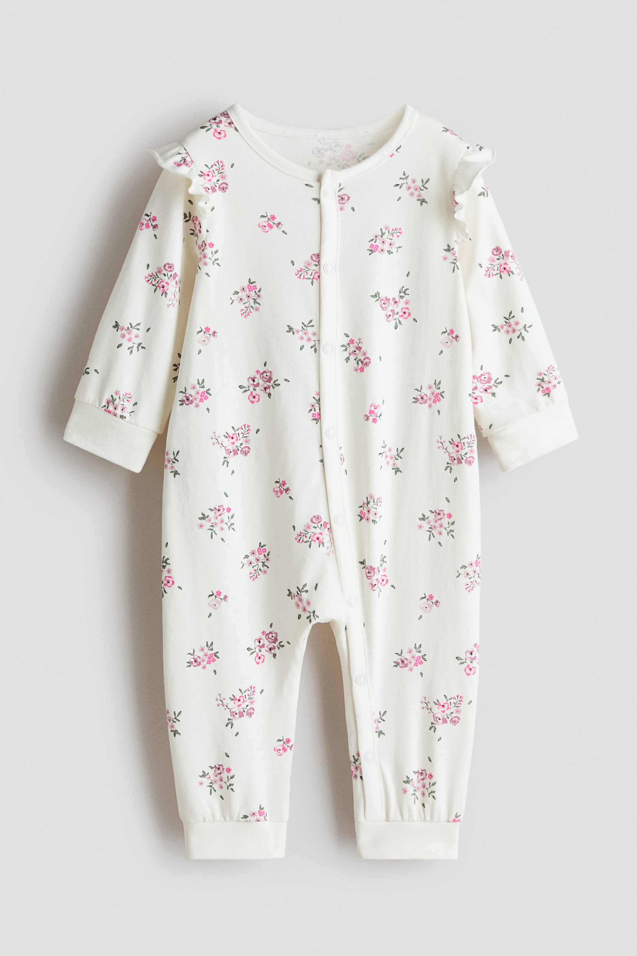 View larger image: Shown from the front, the sleepsuit is made from off-white jersey fabric with an all-over pink floral print, long-sleeved with ruffled shoulder detailing and full-length legs with cuffed hems. Press-studs run down the front and along one leg.