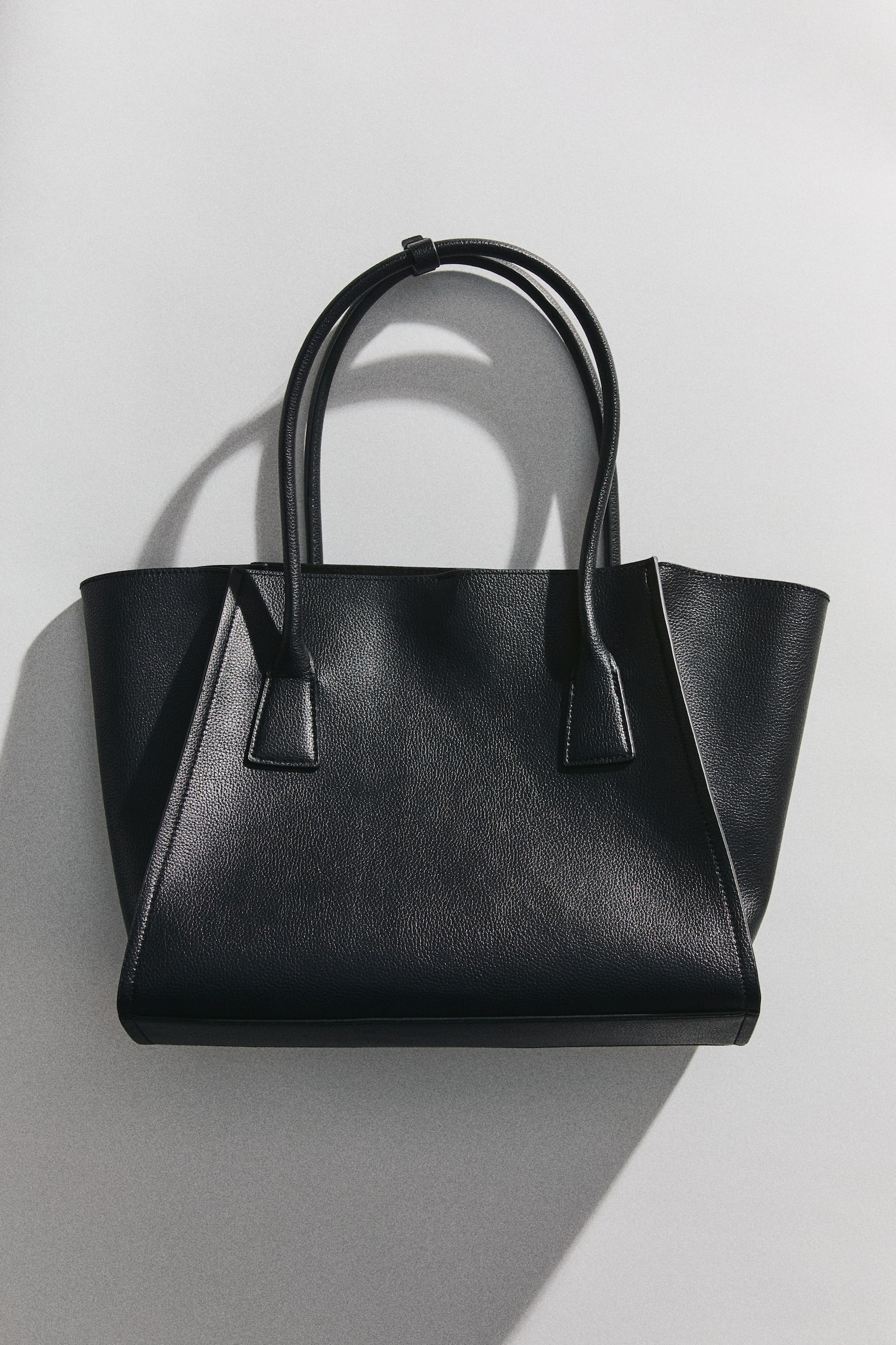 A black tote bag is showcasing its front, featuring a textured material and a structured, tapered shape with two slender top handles joined by a small strap. - 1