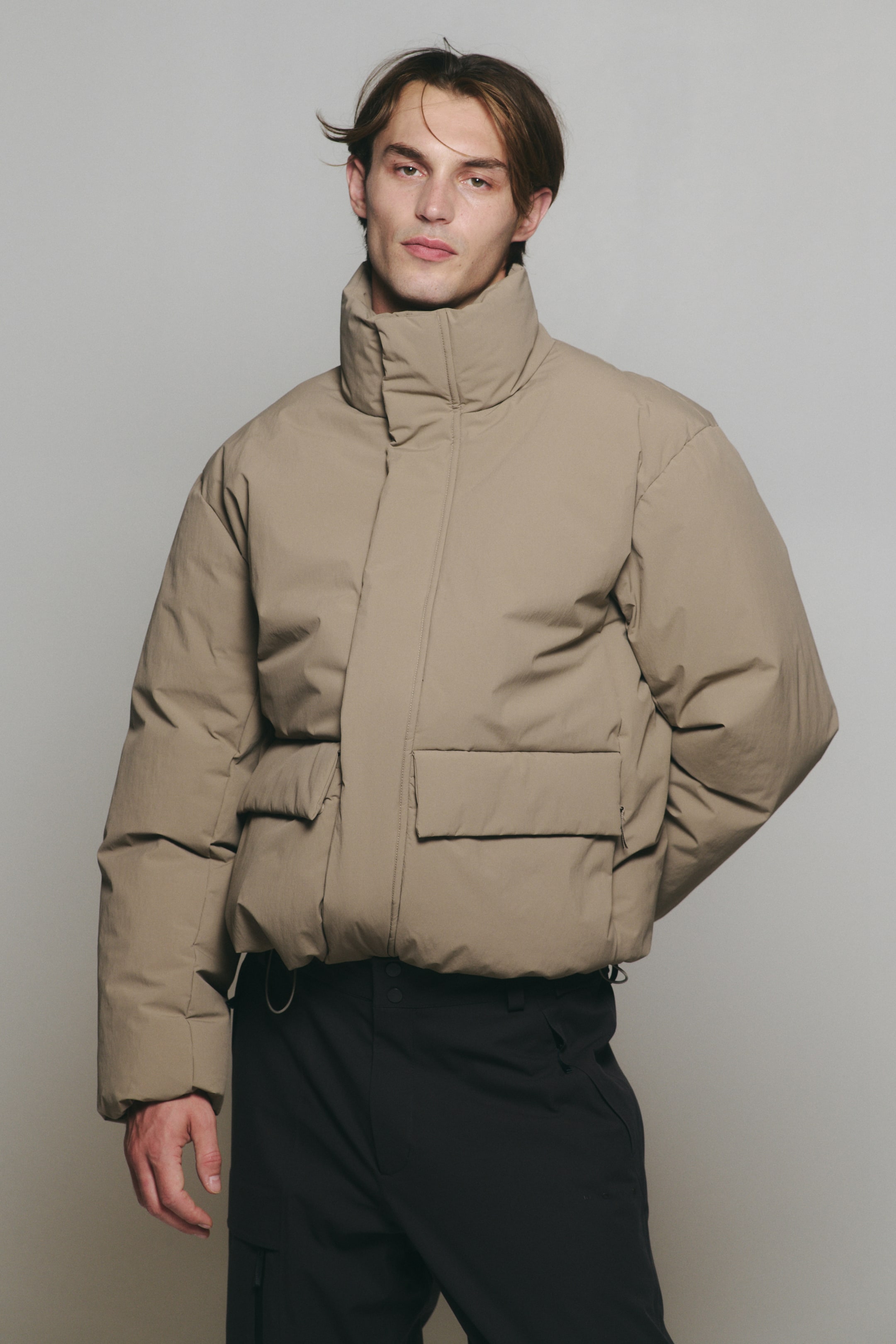 View larger image: A beige puffer jacket featuring a high stand-up collar, a concealed front fastening, and large front flap pockets is being worn by the model. The jacket is styled with dark trousers.