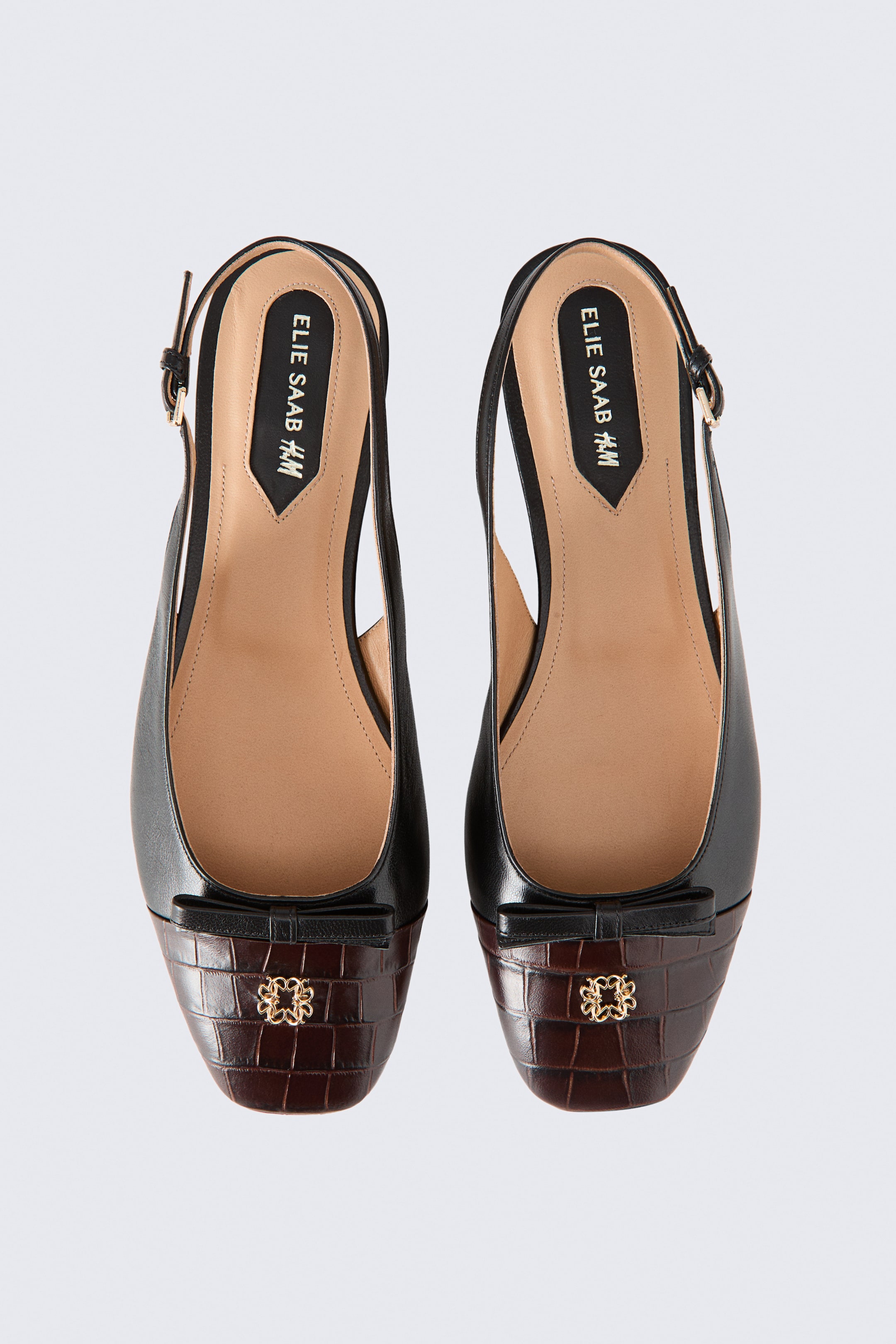 Vis større bilde: Et par Bow Detail Leather Slingbacks viser frem et elegant design i glatt svart skinn med mørkebrune, firkantede tåkapper med krokodillepreg. Hver sko har en liten svart lærsløyfe dekorert med et dekorativt emblem i gulltone på tåpartiet, samt justerbare slingback-ankelstropper og innersåler i tan.