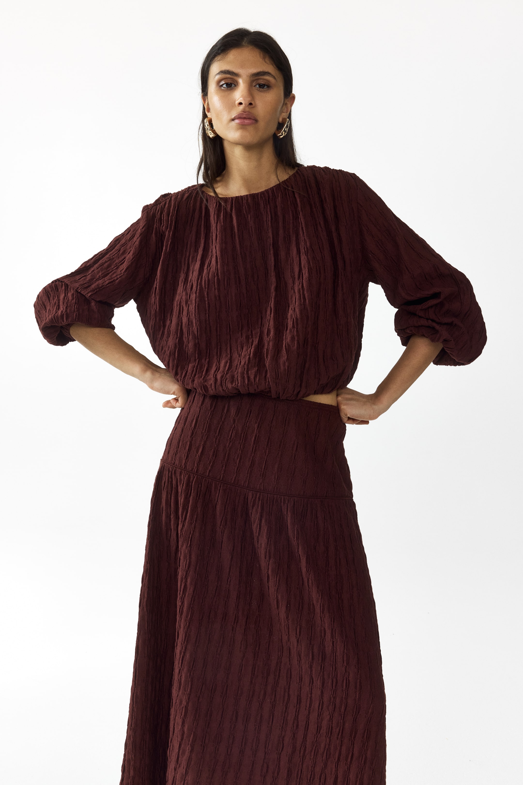 View larger image: A deep reddish-brown two-piece set features a subtle vertical crinkle texture. The top is designed with a round neckline, long gathered sleeves, and an elasticated blouson hem, while the matching midi skirt presents a flat waistband and a flowing silhouette.