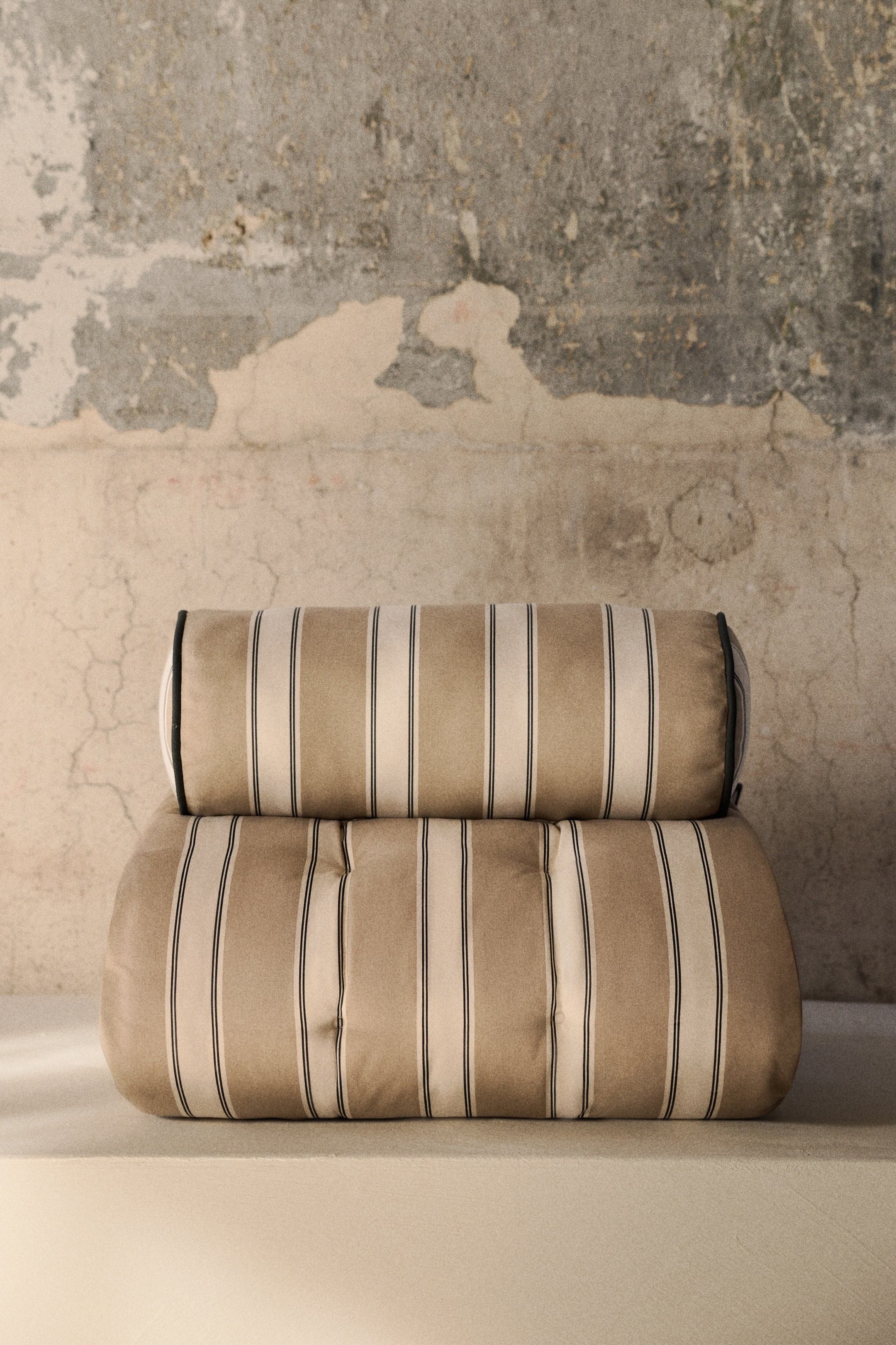 Voir une image plus grande: Un coussin cylindrique repose sur un coussin rectangulaire, tous deux orientés vers l'avant et ornés de rayures verticales en beige clair et blanc cassé, rehaussés de fines lignes noires. Le coussin cylindrique présente un passepoil noir à ses extrémités, tandis que le coussin rectangulaire affiche des indentations capitonnées.