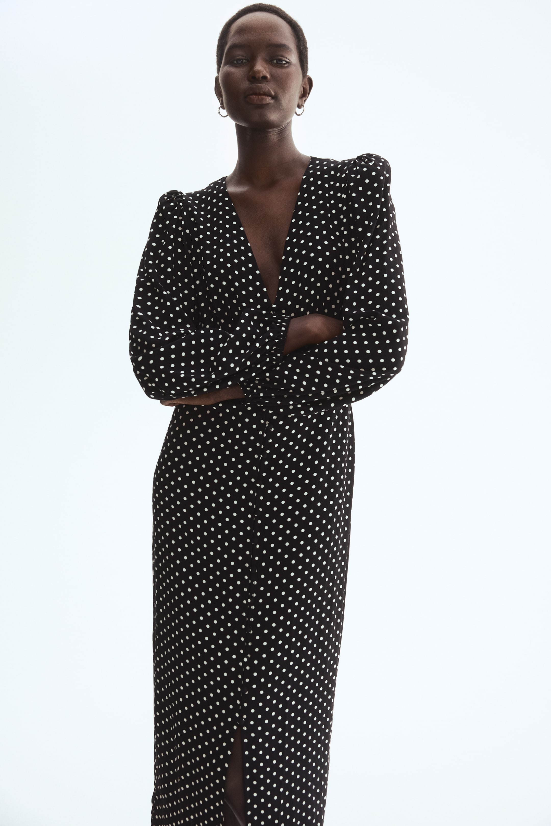הצגת תמונה גדולה יותר: שמלה באורך מידי בצבע שחור עם נקודות לבנות, עם מחשוף V עמוק, כפתורים מכוסים לאורך החזית, ושרוולים ארוכים עם תפרים צרים וגמישים, נלבשת על ידי הדוגמנית. הדוגמנית עומדת עם ידיים שלובות.