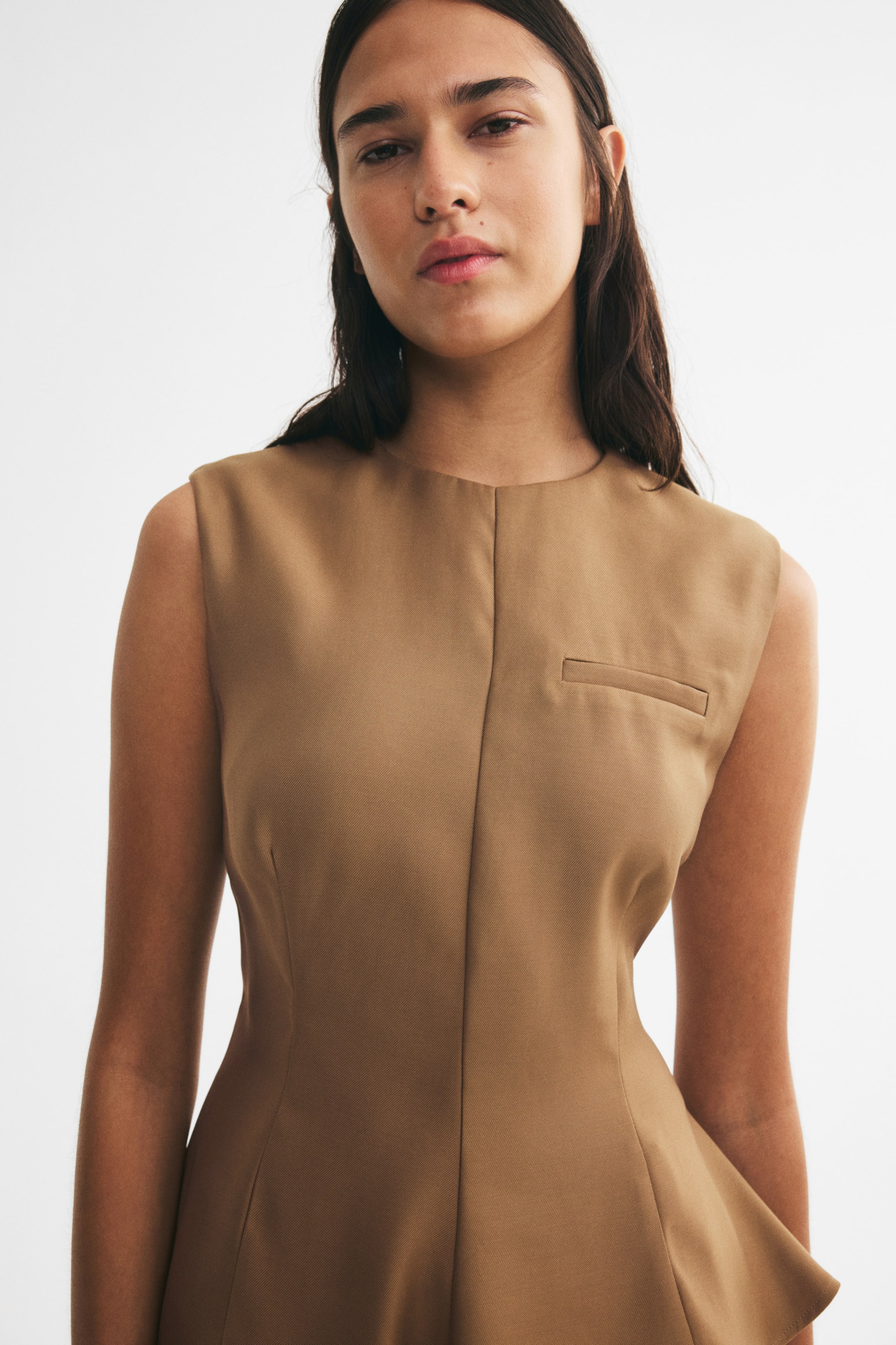 View larger image: The model is wearing a sleeveless tan peplum top. This slim-fit top features a round neckline, a prominent vertical seam running down the center front, a mock welt pocket at the chest, and a flared peplum hem.