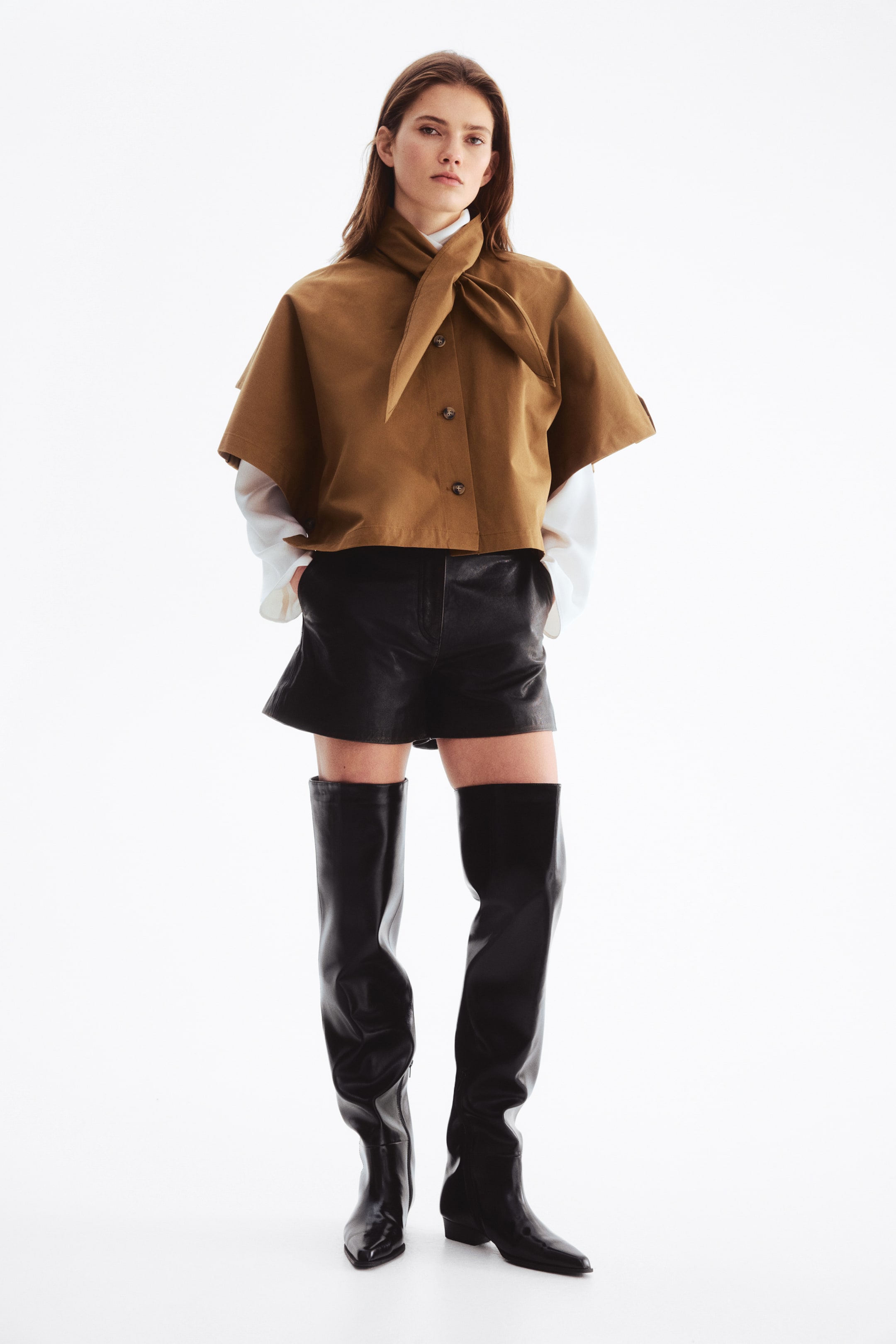 Zobraziť väčší obrázok: Modelka má na sebe krátky plášť v štýle trenčkotu z bavlneného kepru. Plášť je navrhnutý s voľným strihom, rukávmi po lakte a šálovým golierom, ktorý je uviazaný vpredu. Plášť je zapínaný gombíkmi po celej dĺžke vpredu a pod ním je vrstvená biela košeľa s dlhými rukávmi, ktorých rukávy sú viditeľné.