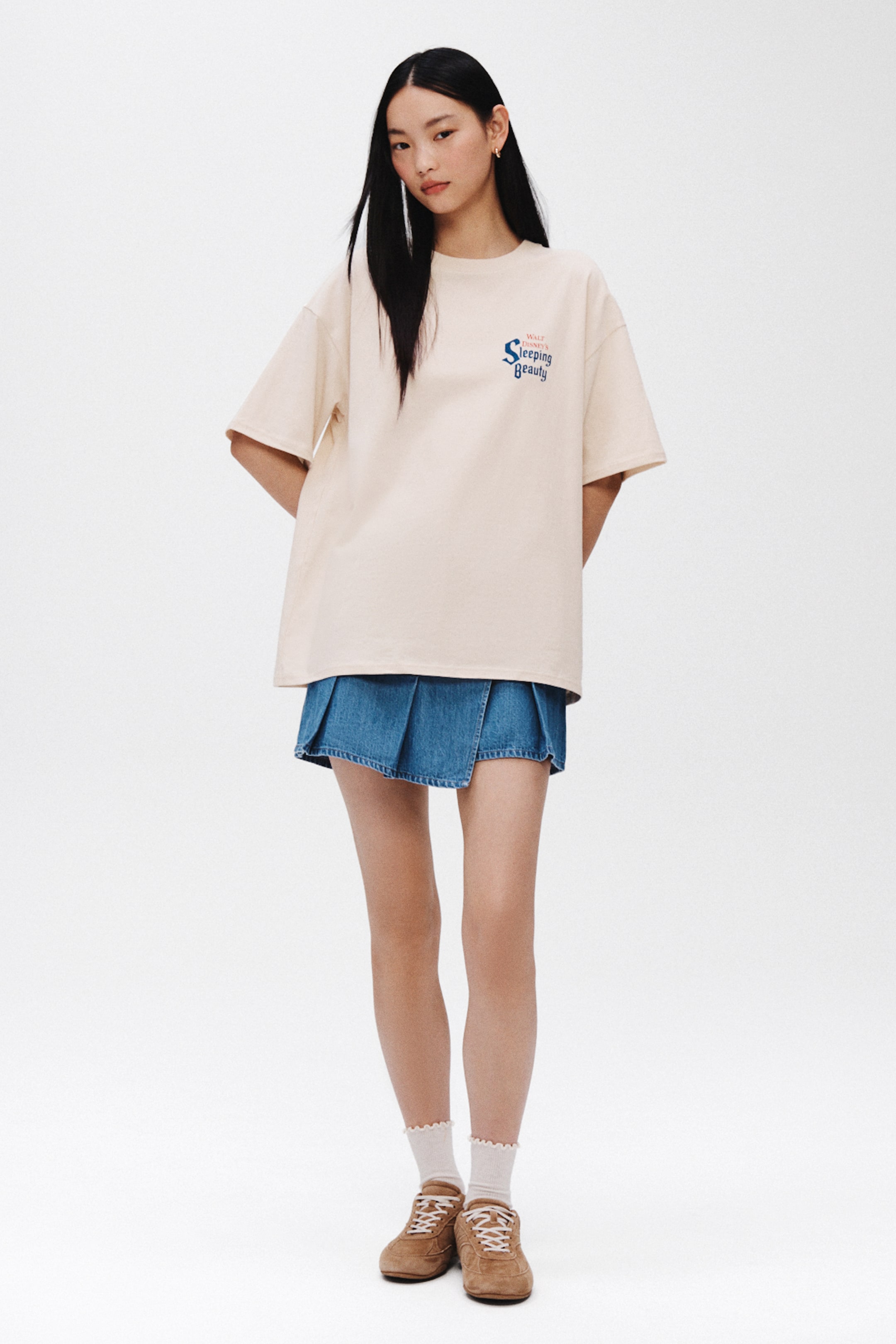 Xem ảnh lớn hơn: Áo phông dáng oversized màu kem, có cổ tròn và tay ngắn, rộng. Một họa tiết màu xanh dương với dòng chữ 'Walt Disney's Sleeping Beauty' xuất hiện ở ngực trái.