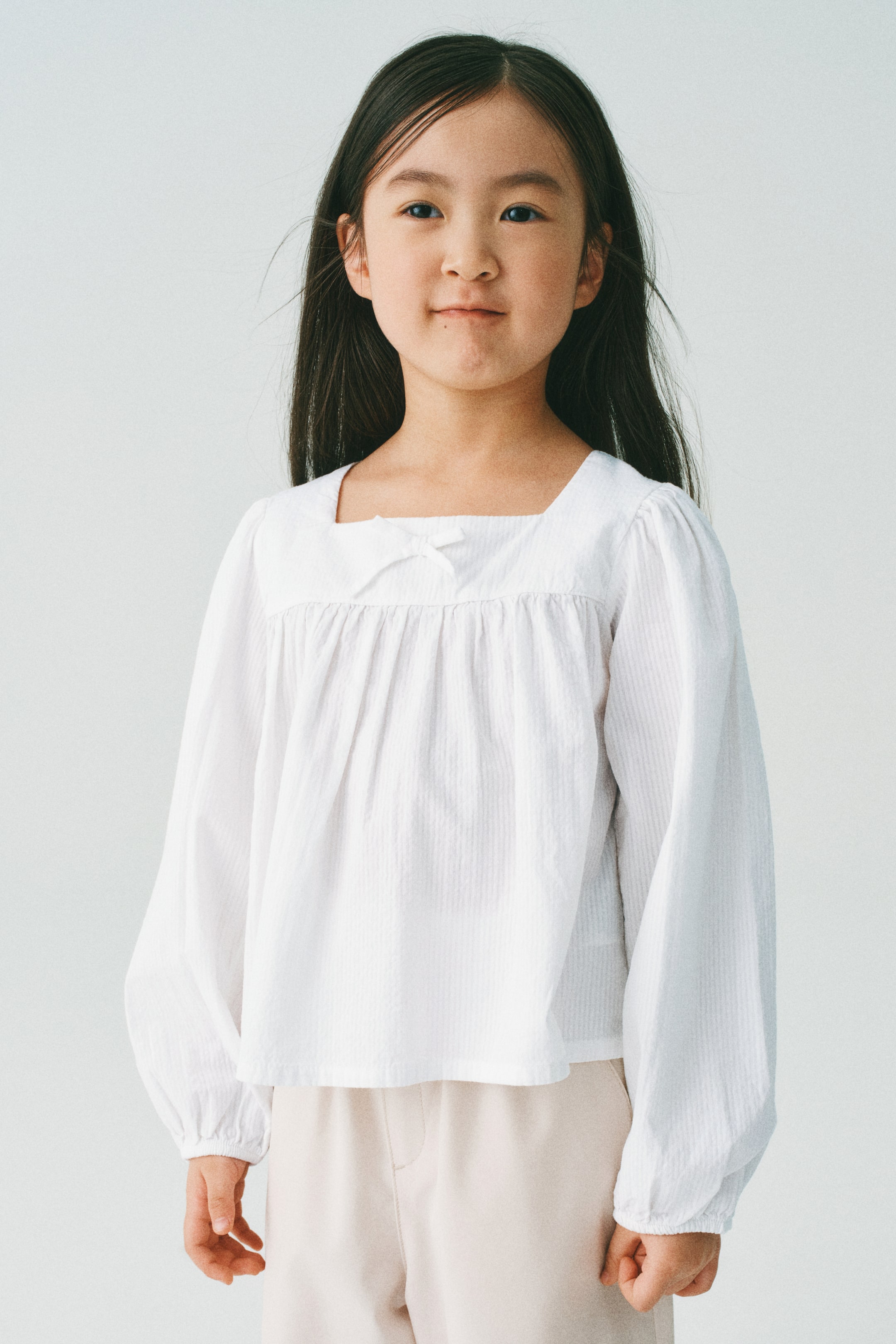 拡大画像を表示: 白い長袖トップスは、微妙なストライプの質感があり、スクエアネックライン、中央前部の小さなリボンディテール、そしてギャザー入りの伸縮性カフスが特徴です。ライトベージュのパンツの上に着用されています。