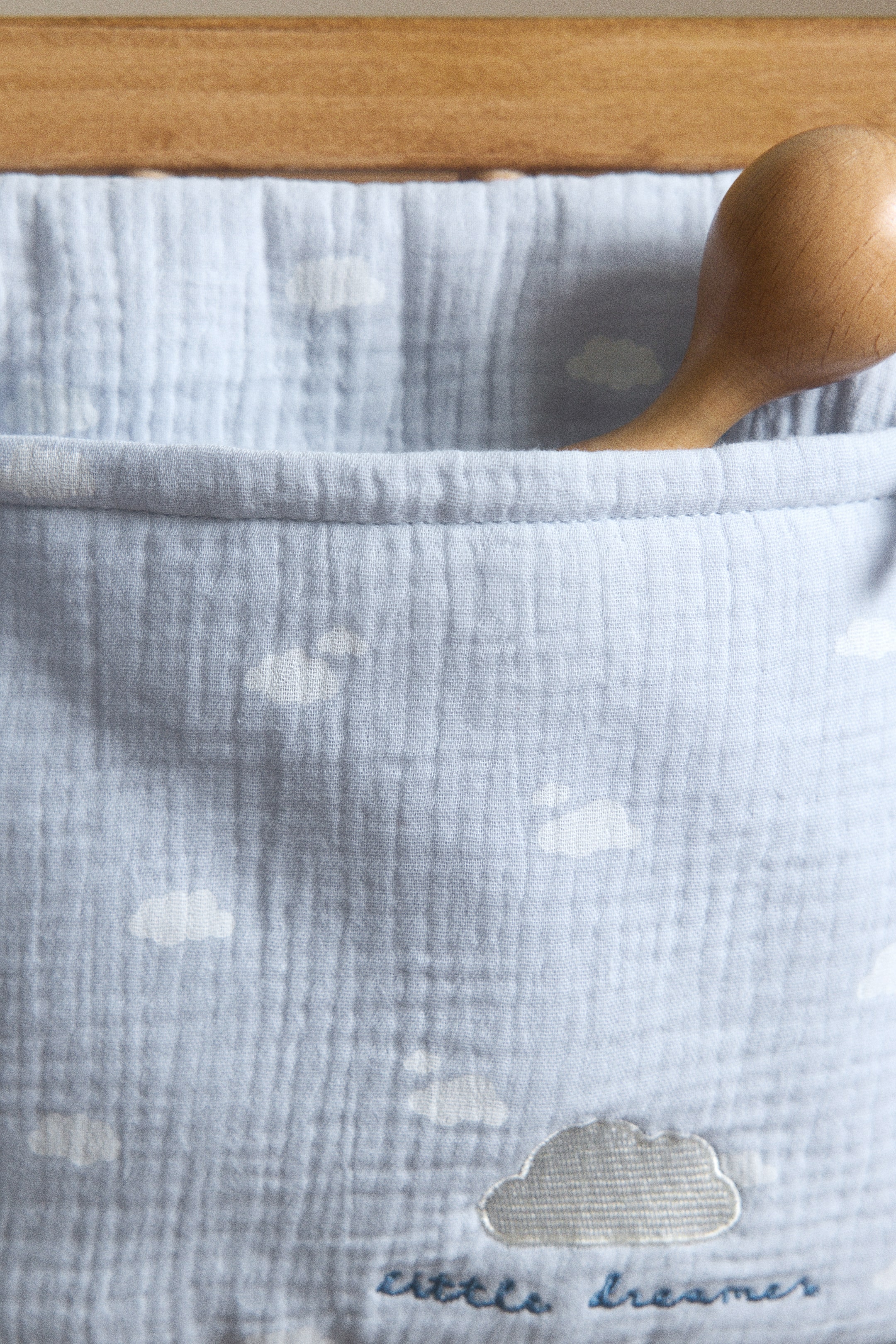 View larger image: A light blue fabric pocket organizer featuring a crinkled texture with white cloud patterns. A gray embroidered cloud and "little dreamer" text detail the lower section, while a wooden object rests inside its open top pocket.