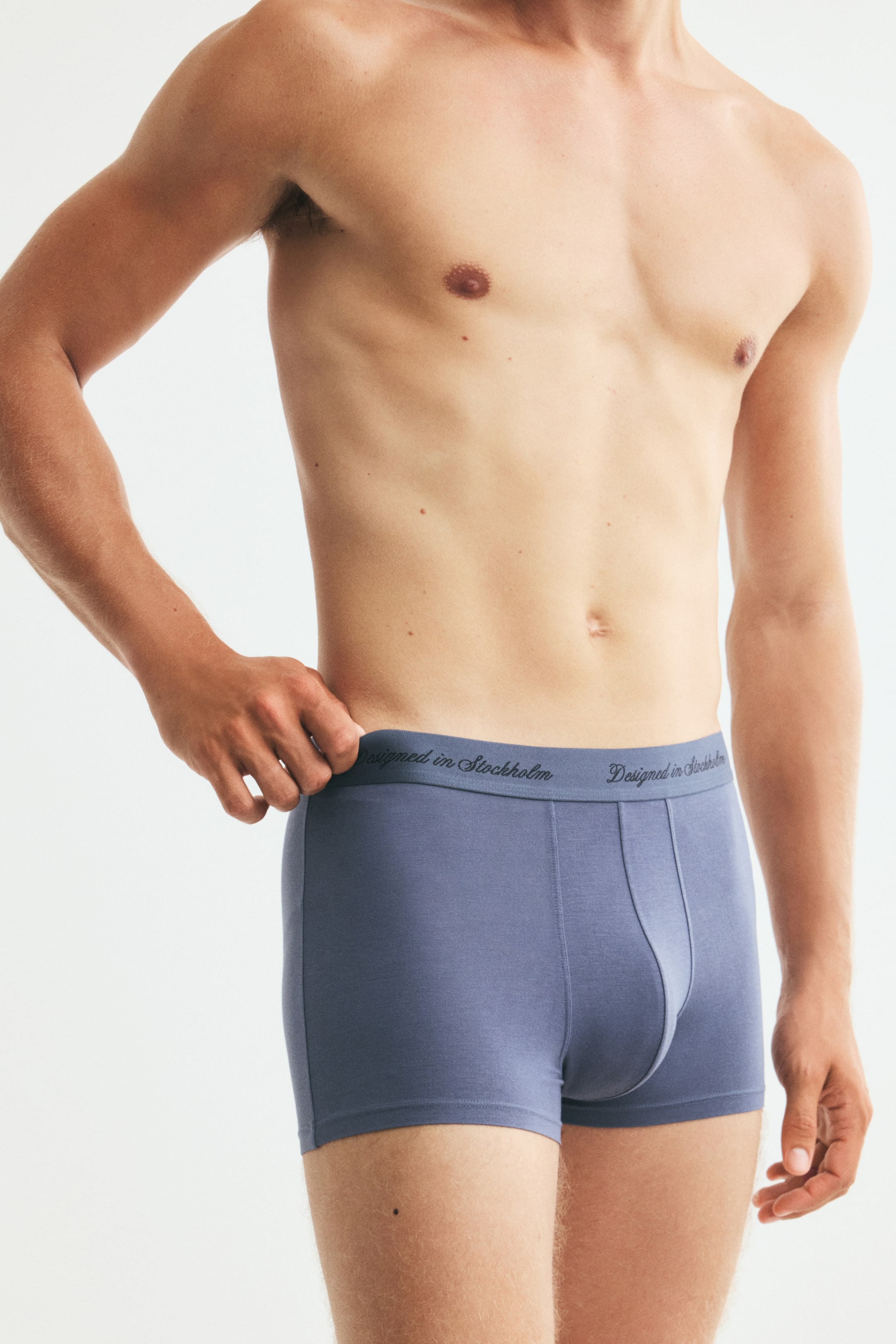 View larger image: The model is wearing blue-grey stretch jersey trunks with short legs and flatlock seams. The model is lightly holding the elastic waistband, which features the text "Designed in Stockholm" across it.