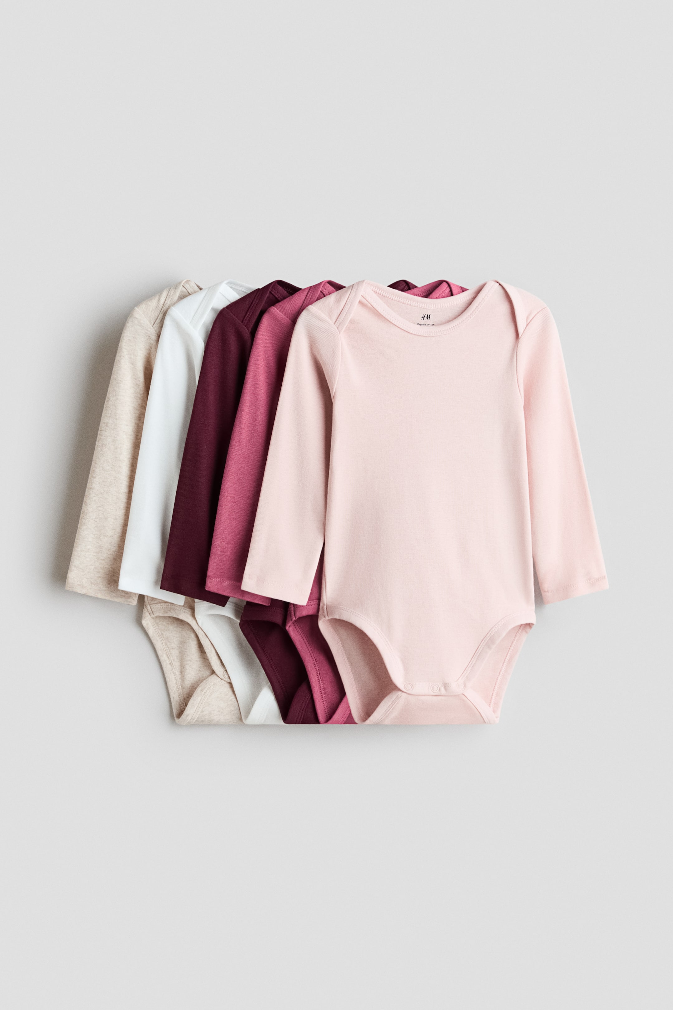 View larger image: Long-sleeved bodysuits crafted from soft cotton jersey are showcased in a neatly fanned stack, with the front facing forward. These bodysuits feature envelope necks for hassle-free dressing and press-studs at the bottom for convenient changing. Available in a variety of colours, including light beige, white, burgundy, and pink.