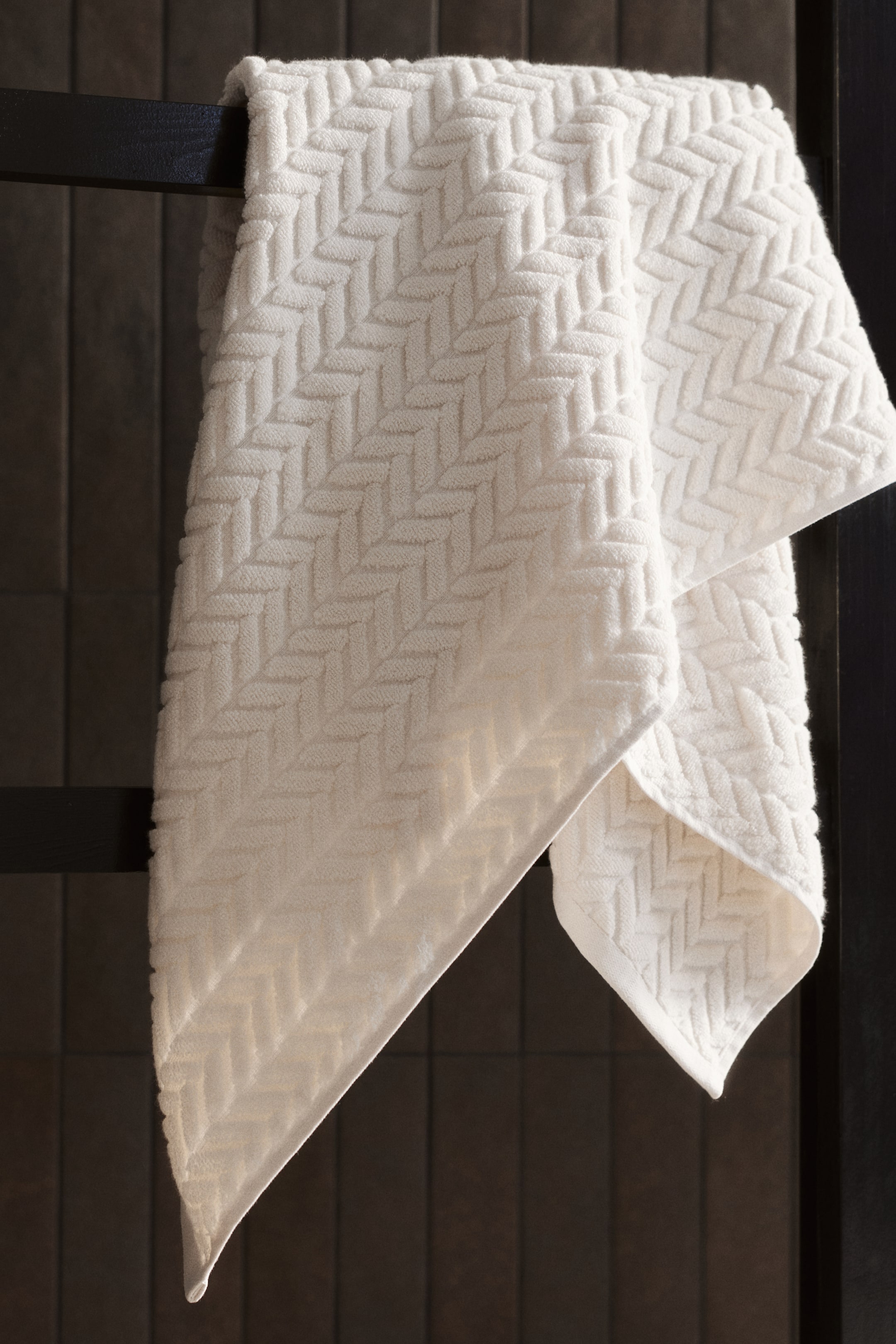 View larger image: A cream-coloured bath towel is showcasing a textured, cable knit-inspired design across its soft cotton terry material while draped over a dark bar.