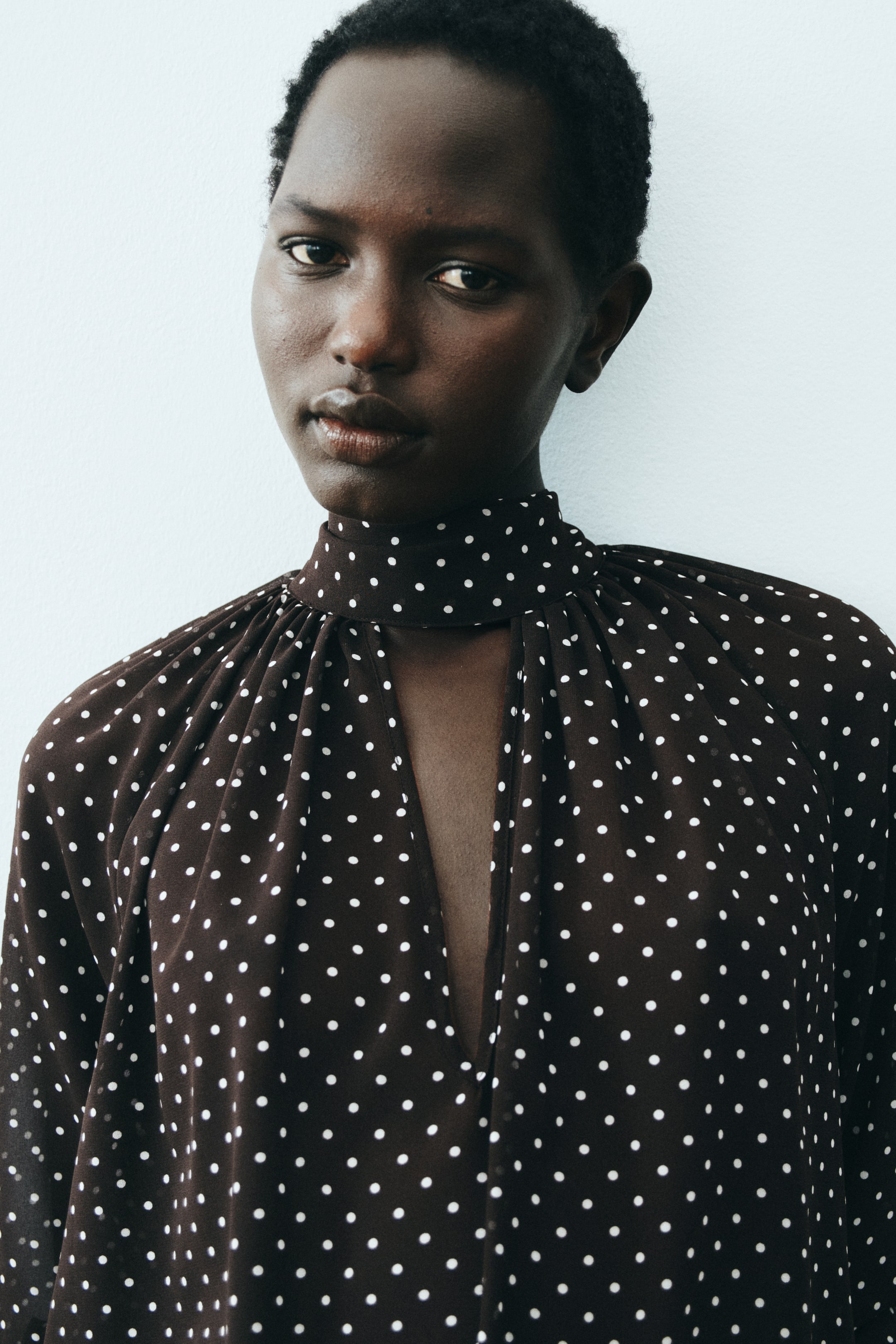 Zobacz większy obraz: Ciemnobrązowa bluzka w białe grochy z wysokim stojącym kołnierzem, który przechodzi w głęboki dekolt w kształcie litery V, oraz marszczonym materiałem na ramionach.