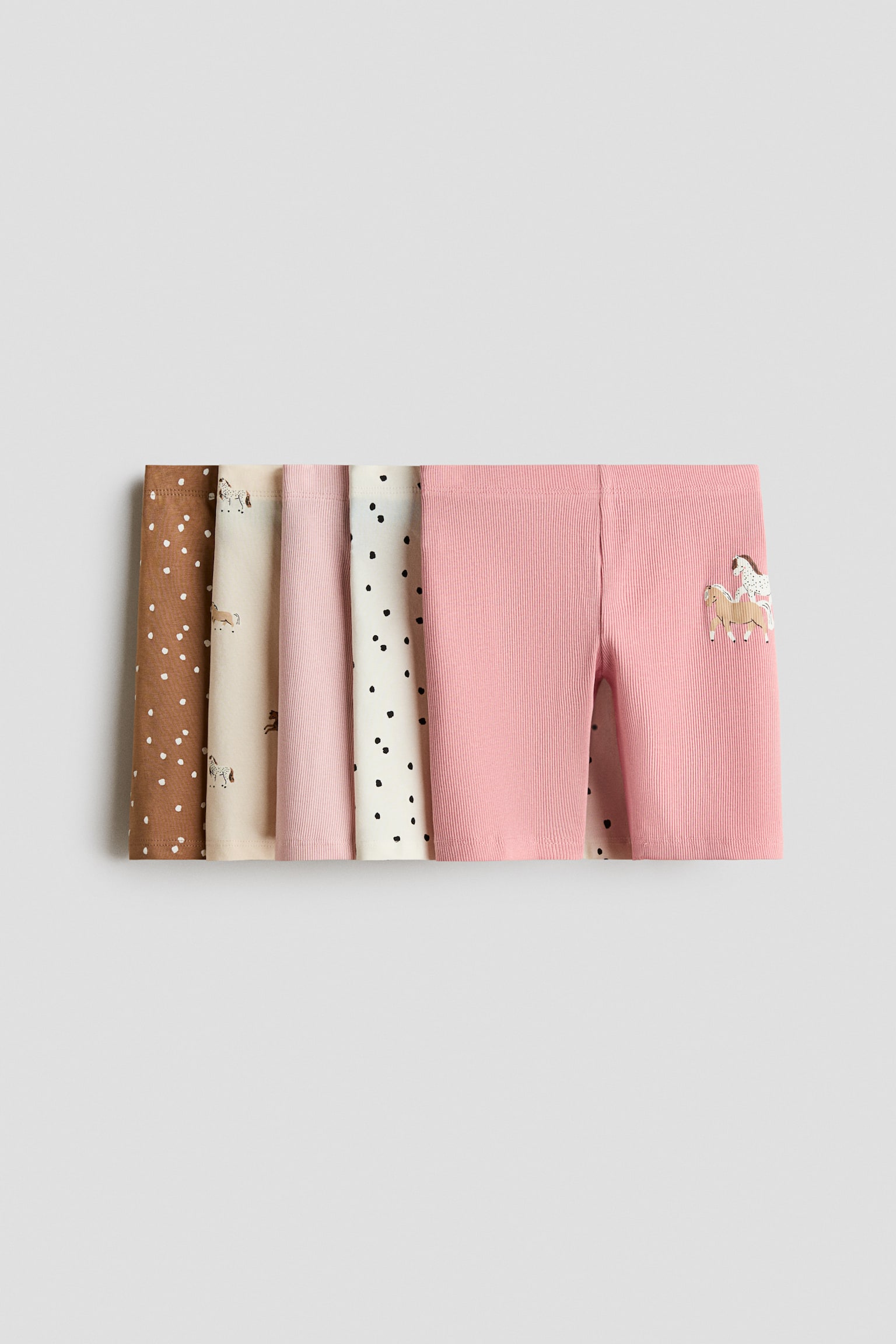 A set of five pairs of ribbed knit shorts is lying flat in an overlapping row, displaying a variety of colors and patterns. The front-facing pair is a solid pink with a small horse graphic on the lower leg, while the other pairs layered behind are featuring brown with white dots, beige with horse illustrations, solid light pink, and off-white with black dots.