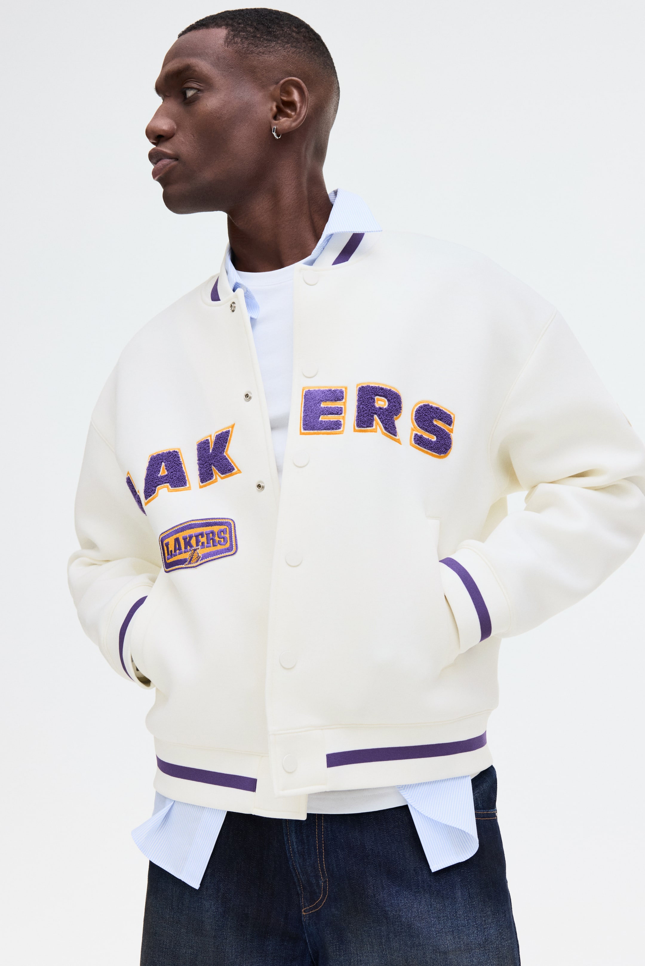 View larger image: A cream varsity jacket showcases purple and orange chenille 'LAKERS' lettering across the chest, along with a smaller 'LAKERS' logo patch. This jacket features a ribbed collar, cuffs, and hem, detailed with purple and yellow stripes, and closes at the front with snap buttons. It is worn over a light blue striped shirt and a white t-shirt.