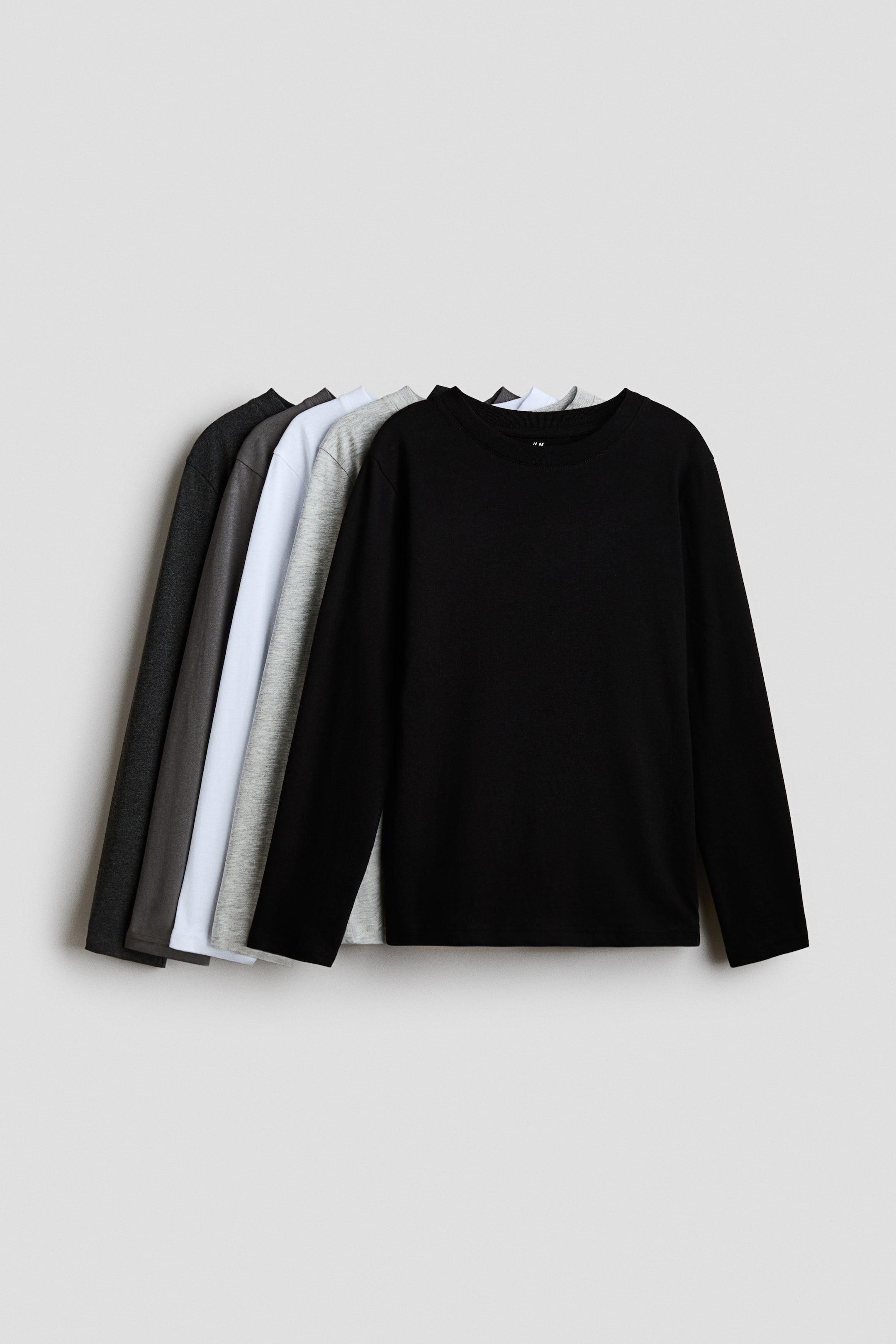 View larger image: Long-sleeved T-shirts in soft cotton jersey are displayed in a fanned stack, with the black shirt positioned at the front and facing forward. Each shirt features a round, rib-trimmed neckline and long sleeves, appearing in solid colours of black, light grey, white, and various shades of dark grey.