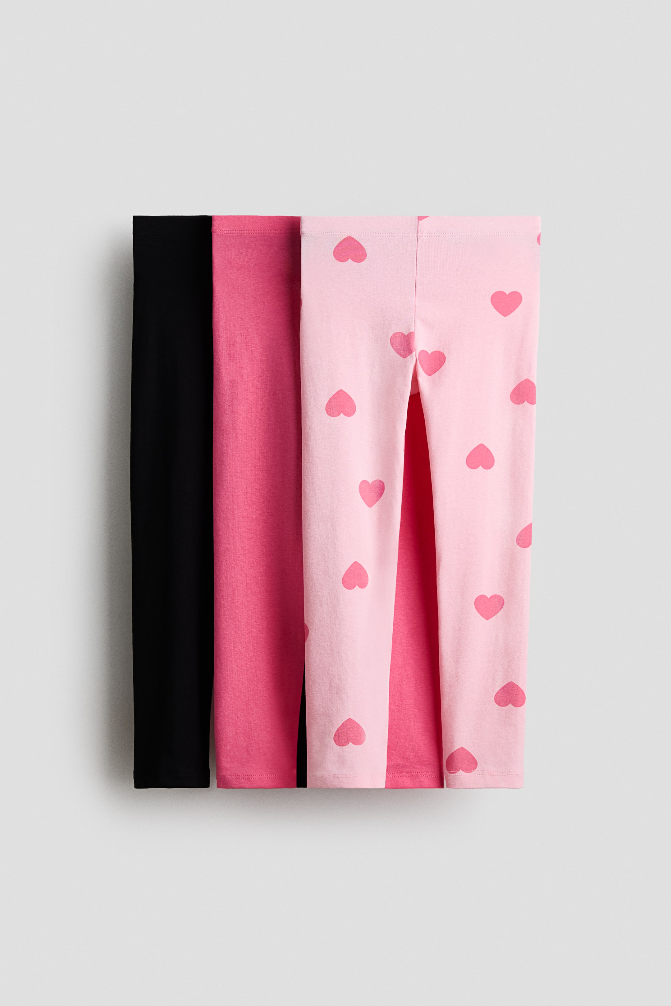 Größeres Bild ansehen: Drei Paar Leggings, aus weichem Baumwolljersey gefertigt und mit einem elastischen Bund ausgestattet, sind frontal abgebildet. Die Designs sind in einfarbigem Schwarz, einfarbigem Fuchsia und Hellrosa mit einem durchgehenden, dunkleren rosa Herzmuster gehalten.