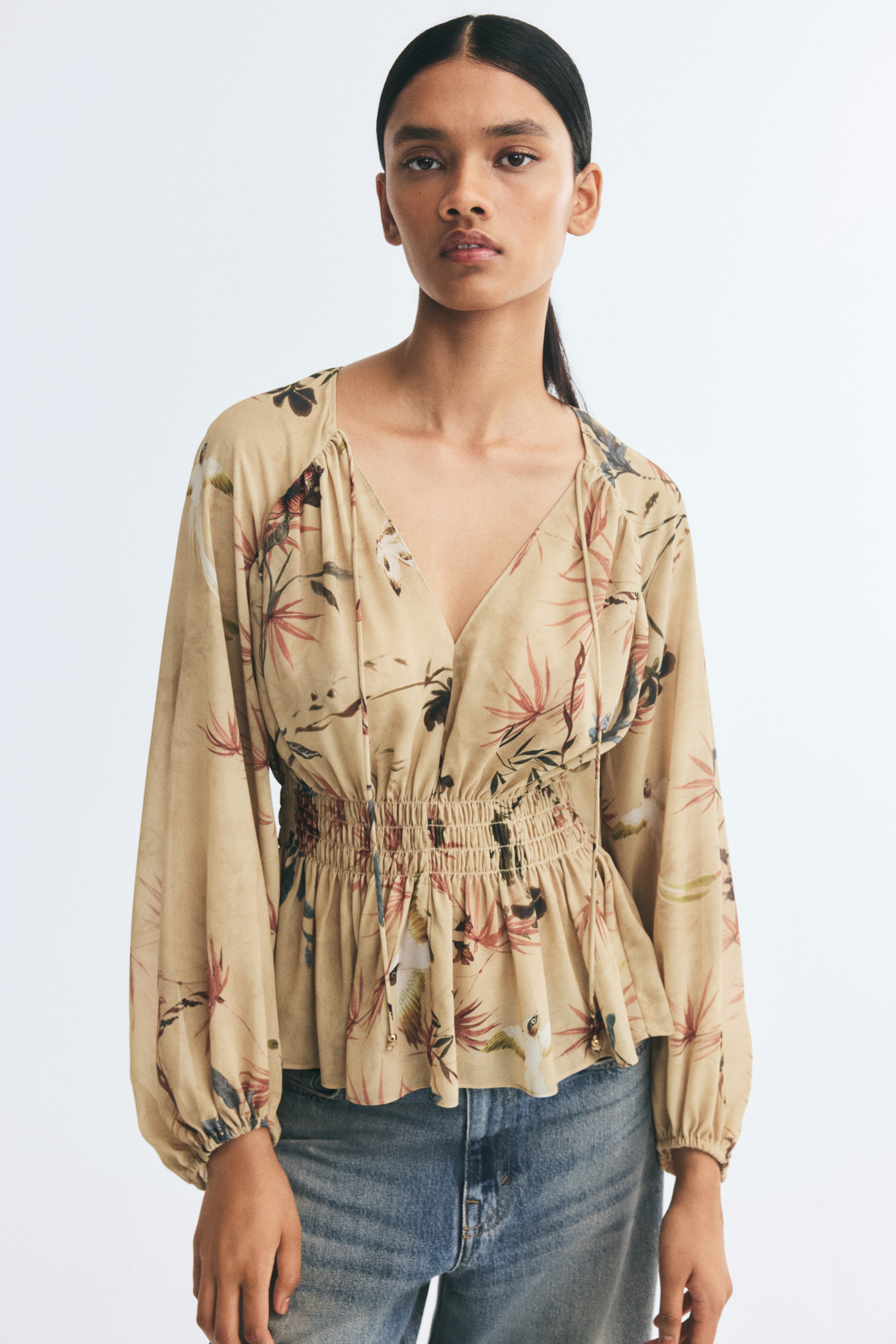 Visualizza immagine più grande: La modella indossa una blusa beige con stampa floreale, caratterizzata da un profondo scollo a V con sottili lacci pendenti, maniche lunghe a palloncino con polsini elasticizzati e un ampio punto smock in vita. La blusa è abbinata a jeans in denim blu.