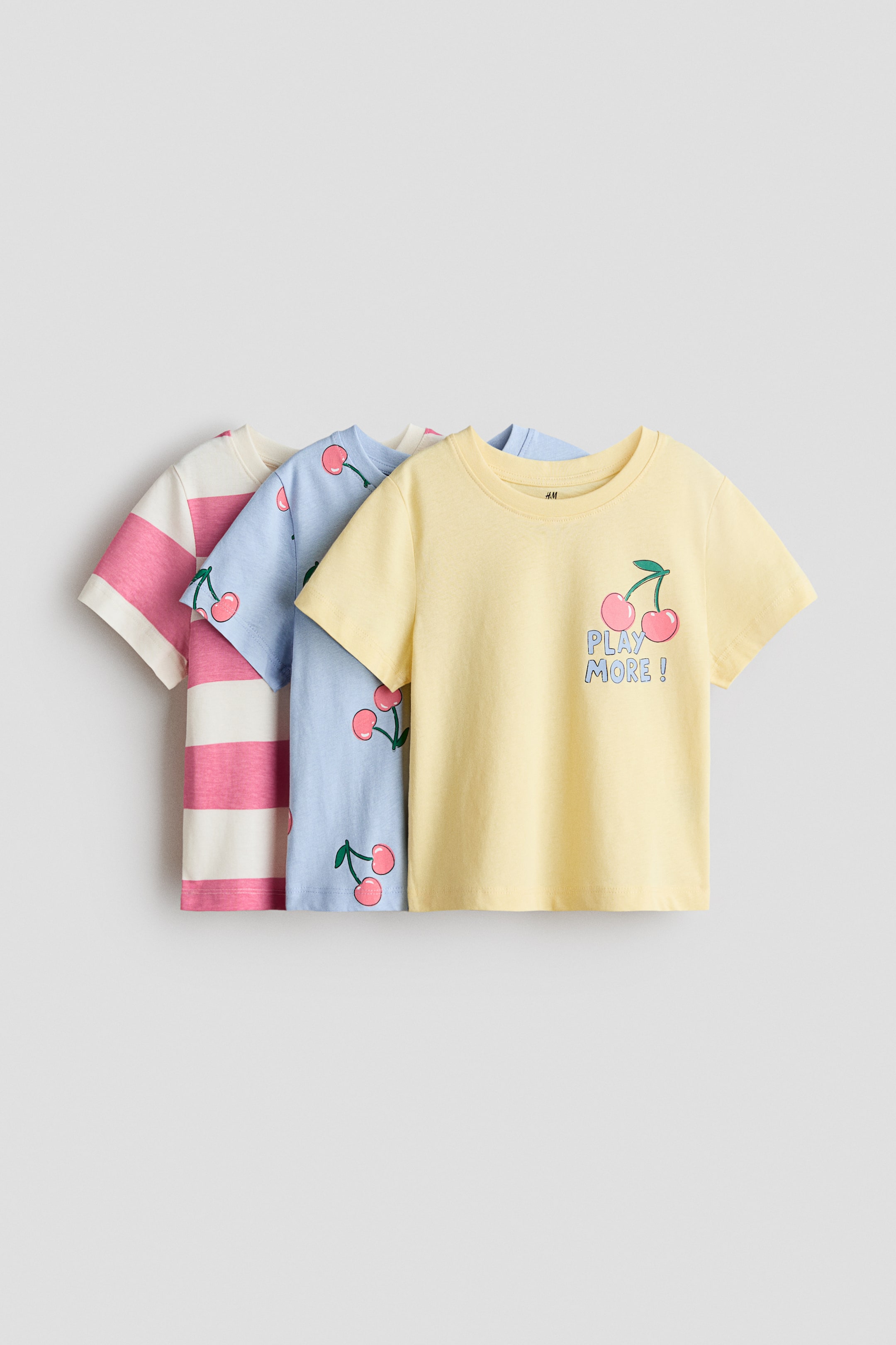 View larger image: Three children’s cotton jersey t-shirts are shown facing front in a layered arrangement. The front t-shirt is light yellow featuring a cherry graphic and "PLAY MORE!" printed on the chest, the middle is light blue displaying an all-over cherry pattern, and the back t-shirt features pink and white horizontal stripes.