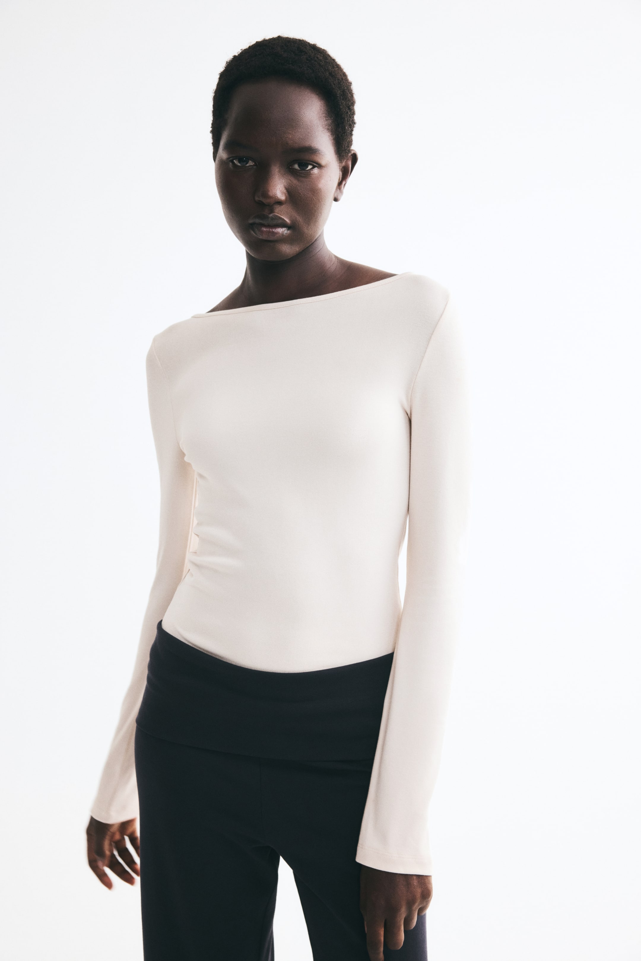 View larger image: The model is wearing a cream-coloured fine-knit top with a delicate boat neckline and long sleeves. The fitted top is paired with black trousers featuring a wide fold-over waistband.