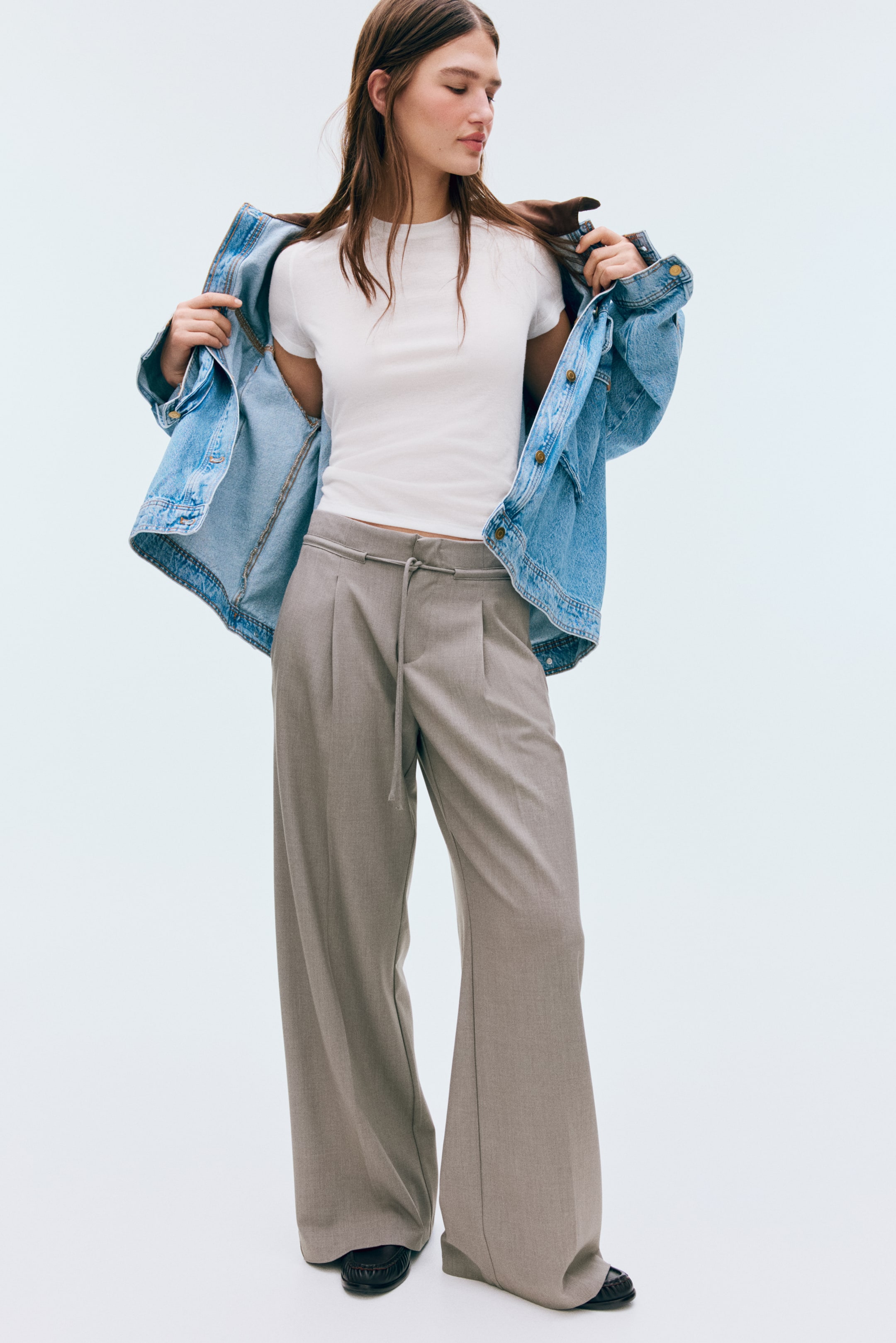 View larger image: The taupe wide-leg trousers feature a relaxed fit and a drawstring waist, with front pleats enhancing their fluid drape. They pool slightly over black shoes, revealing their full-length design.
