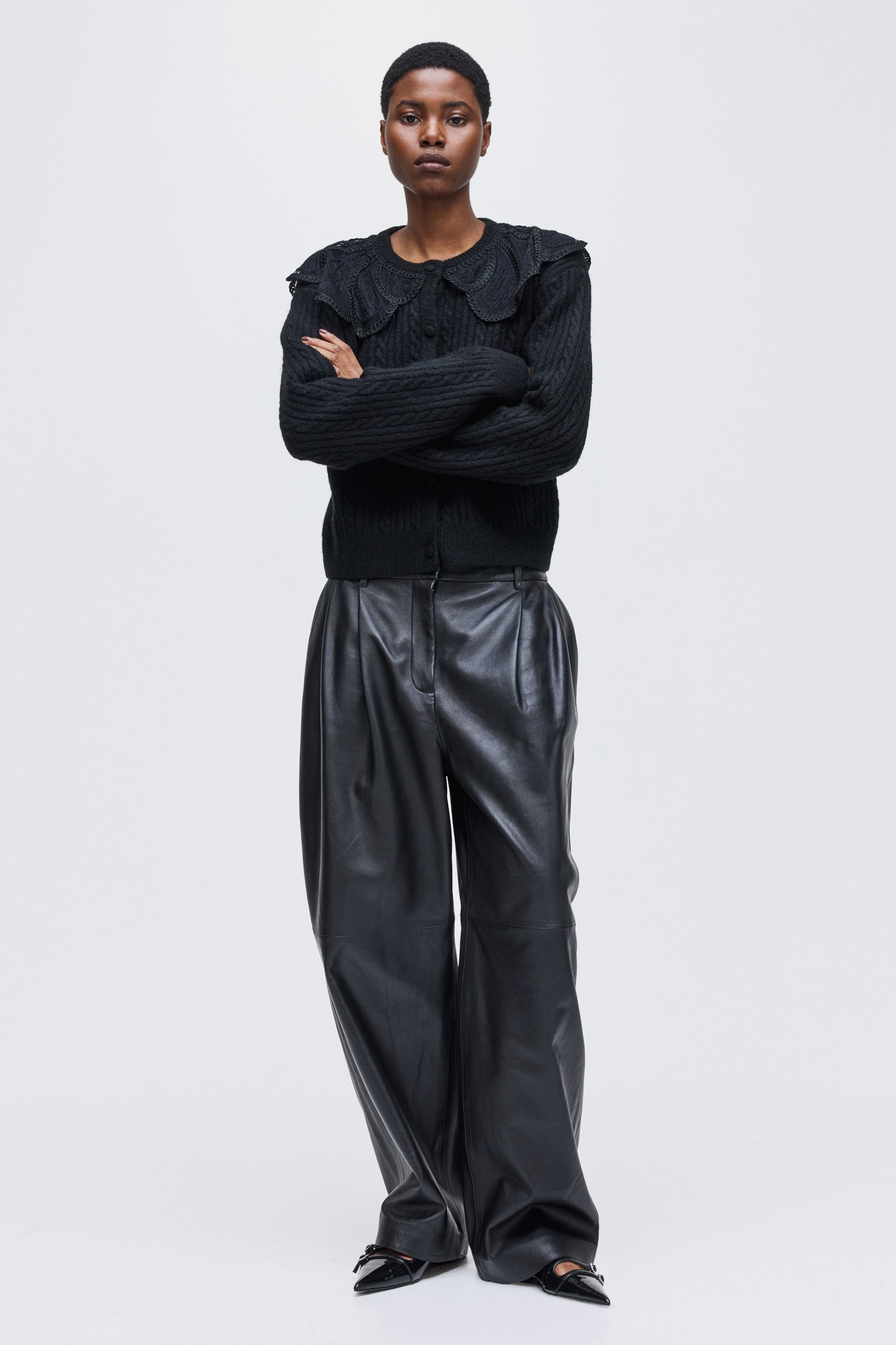 拡大画像を表示: 黒のケーブルニットカーディガンは、大きなスカラップ刺繍の襟とボタンフロントクロージャーが特徴です。それは、高いウエストとフロントプリーツが施された黒のワイドレッグレザーパンツと共に着用されています。