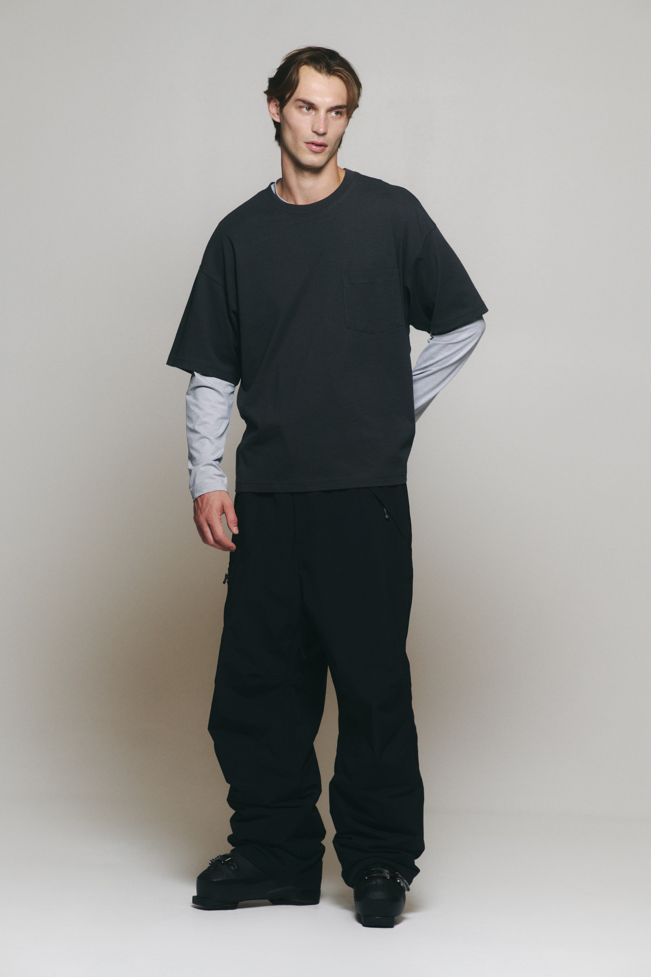 Visualizza l'immagine ingrandita: Una t-shirt girocollo grigio scuro con taschino sul petto è indossata dalla modella, sovrapposta a una maglia a maniche lunghe grigio chiaro. La t-shirt è abbinata a pantaloni neri ampi a gamba larga.