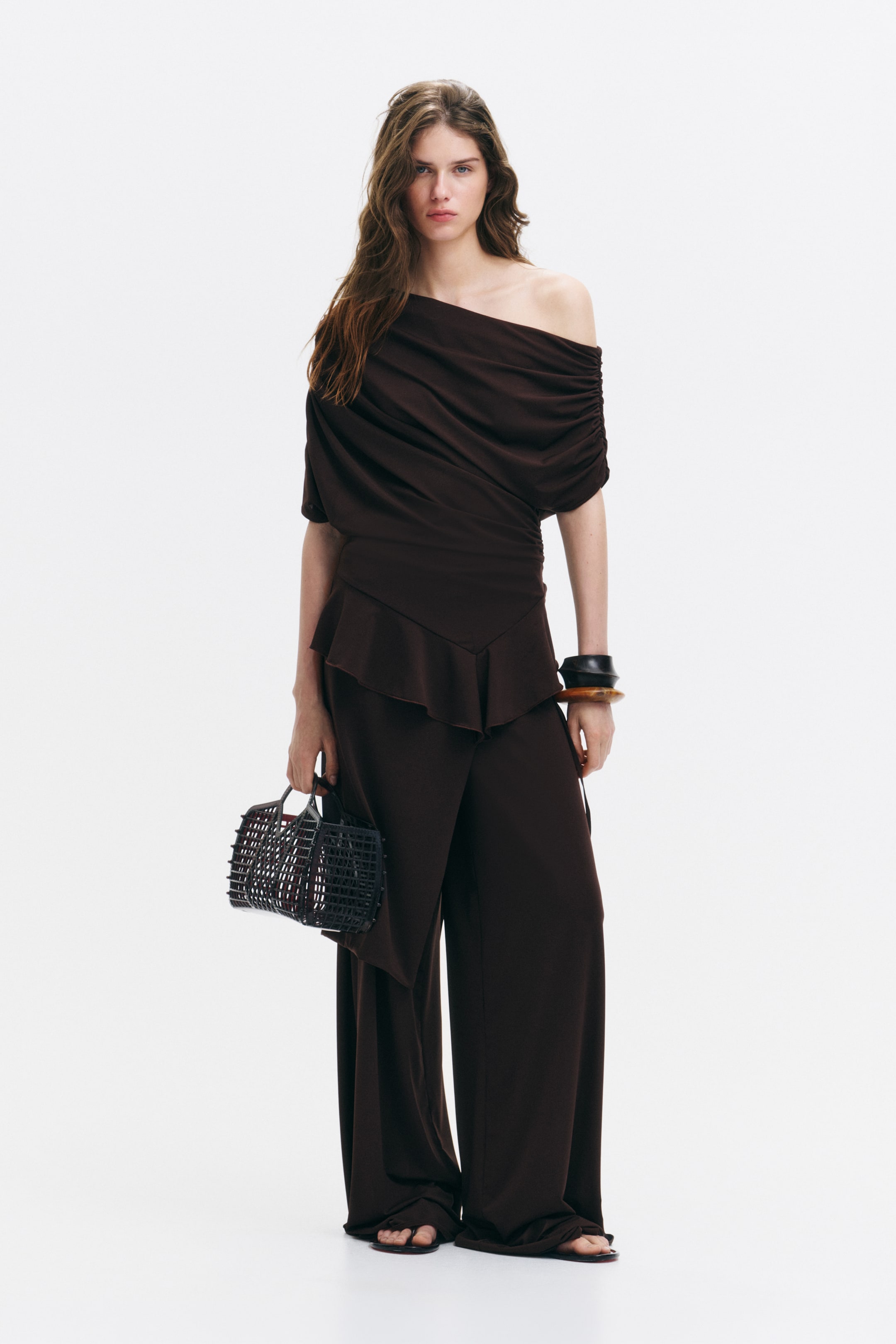 View larger image: The model is wearing a dark brown one-shoulder top crafted from stretch jersey with a subtle sheen. It features gathers throughout, creating a draped effect, an asymmetric neckline, a cap sleeve on one side, a wide sleeve panel around the opposite arm, and an asymmetric, flounced hem. The look is completed with matching wide-leg trousers.