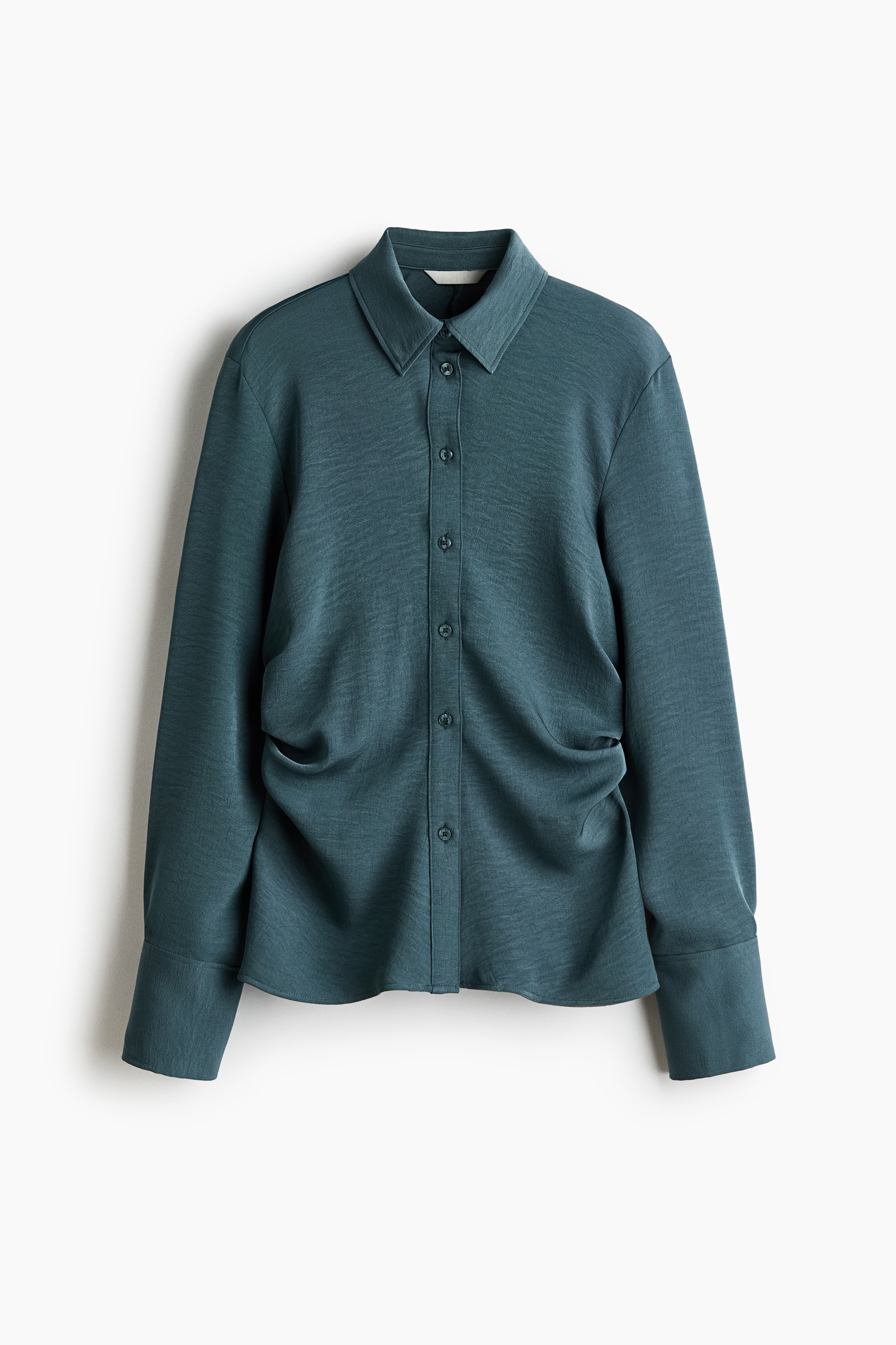 A slate-blue woven shirt with a collar, button-down front, and side gathers is worn by the model. The shirt features long sleeves with wide button cuffs and is styled partially unbuttoned at the straight hem over black trousers.