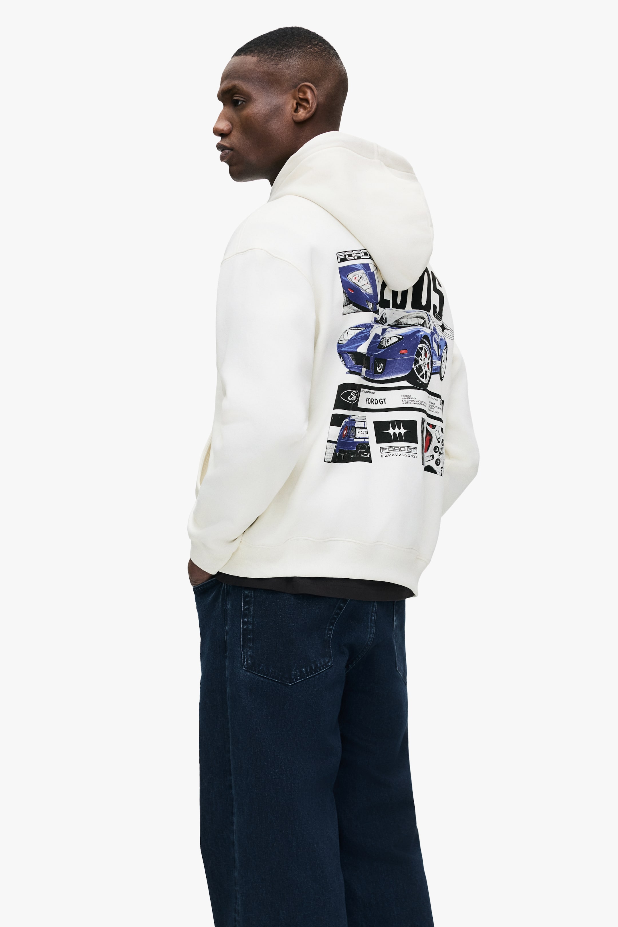 Größeres Bild anzeigen: Ein cremefarbener Hoodie mit einem Grafikdruck von blauen Ford-GT-Autoillustrationen und Text auf der Rückseite wird vom Model getragen. Das Model trägt auch dunkel gewaschene Jeans und ein dunkles Unterhemd unter dem Hoodie.