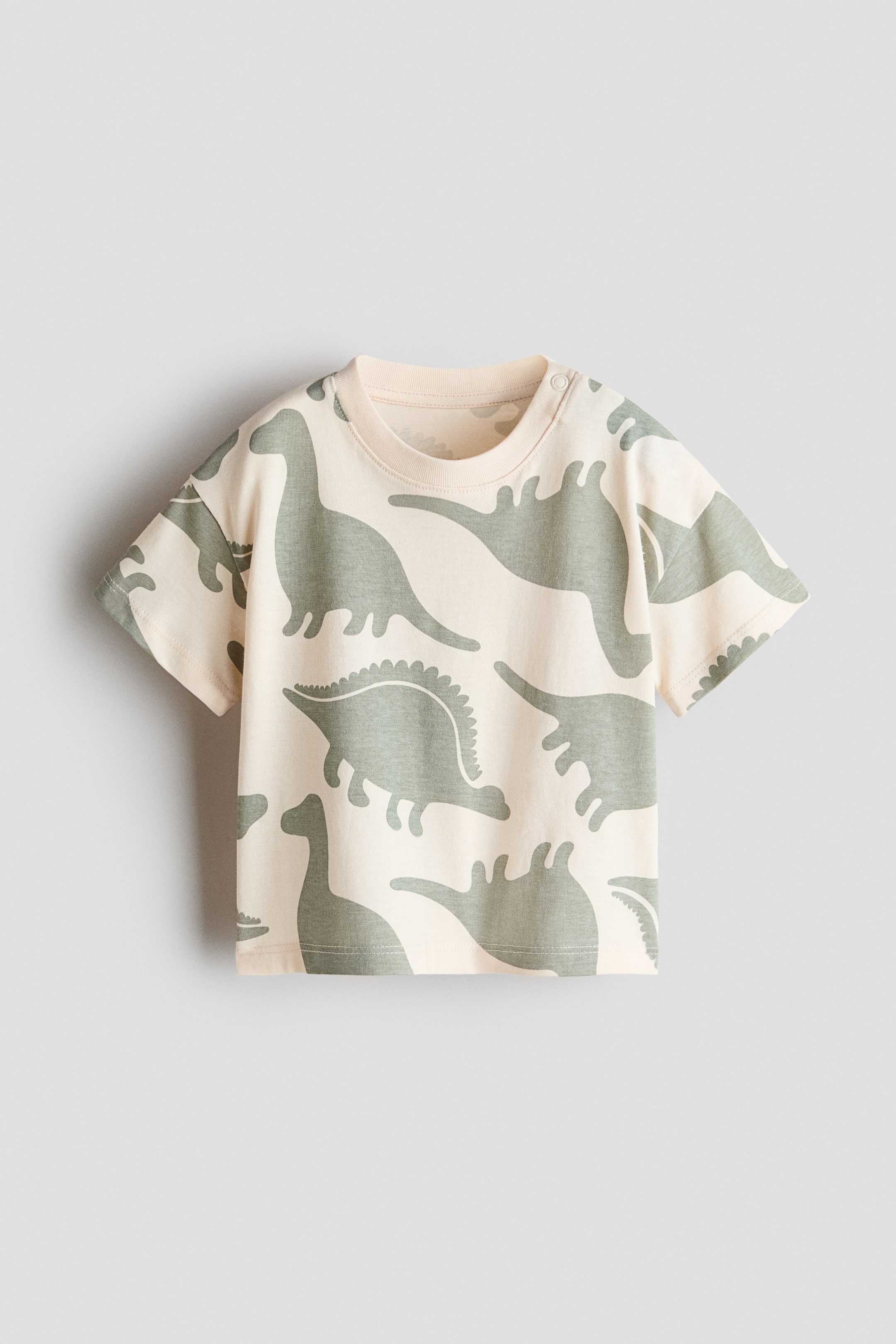 View larger image: Printed cotton T-shirt is lying flat, showcasing its light cream colour and a repeating pattern of greyish-green dinosaurs across the front. It features a ribbed crew neck and two snap buttons on the left shoulder.