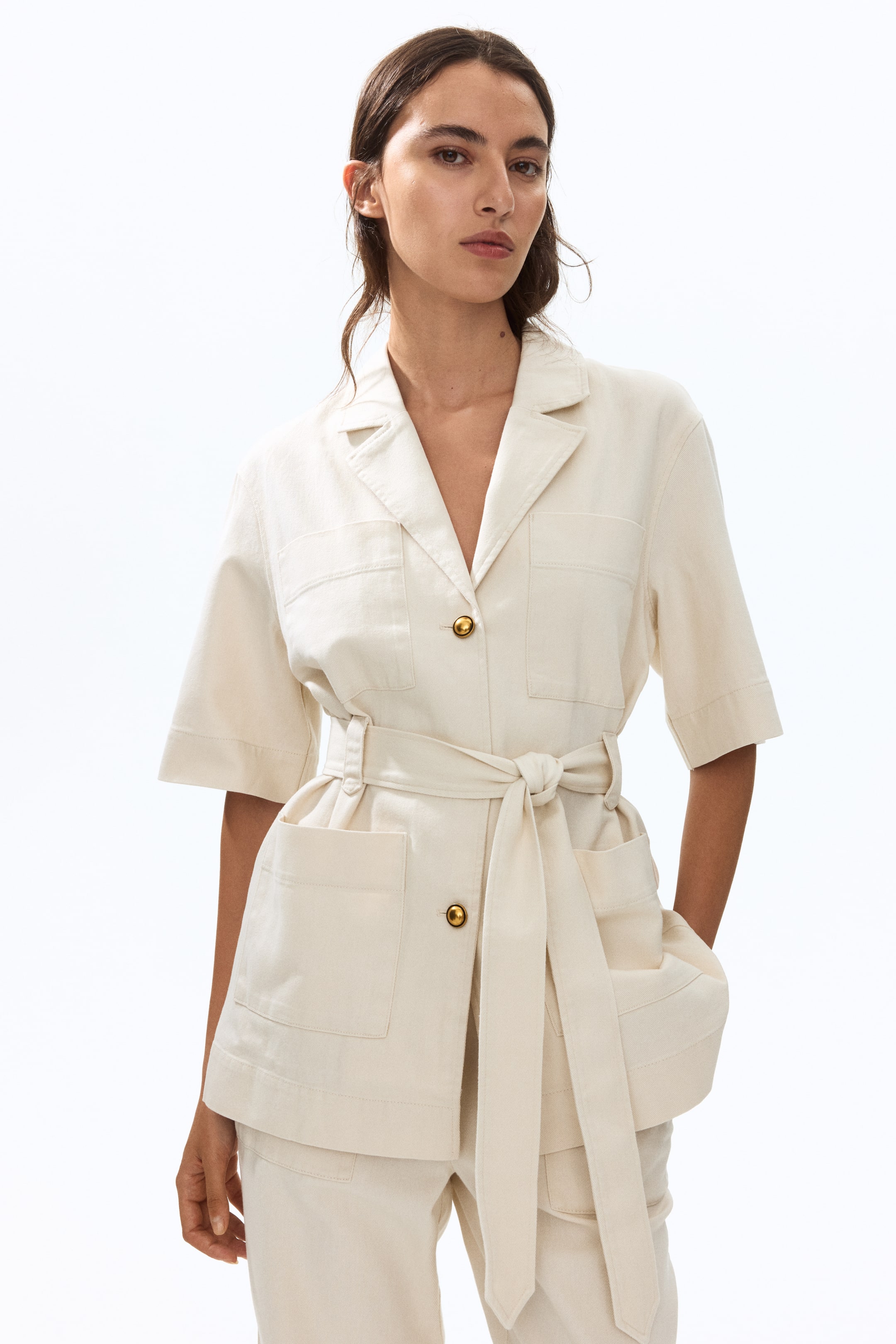 View larger image: A cream-coloured utility jacket featuring notch lapels, short sleeves, and a self-tie belt cinching the waist, presented from a front, slightly angled view. Two chest patch pockets with flaps and two larger patch pockets below the waist are visible, along with gold-toned buttons securing the front closure.