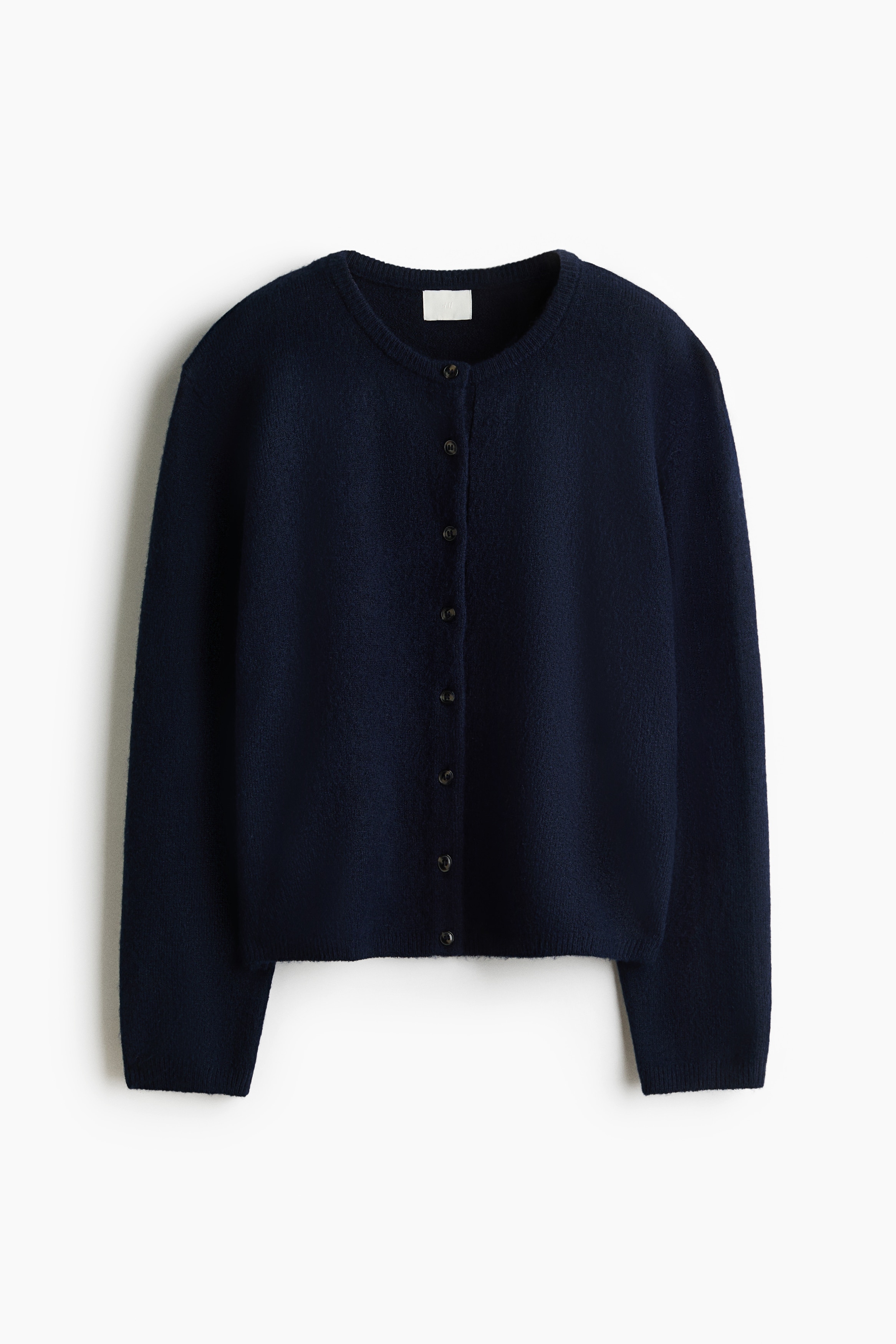 A dark navy blue, loose-fit knit cardigan is worn by the model, featuring a round neckline, button-down front, dropped shoulders, and long sleeves. The cardigan is styled with the top partially unbuttoned and paired with black leather trousers.