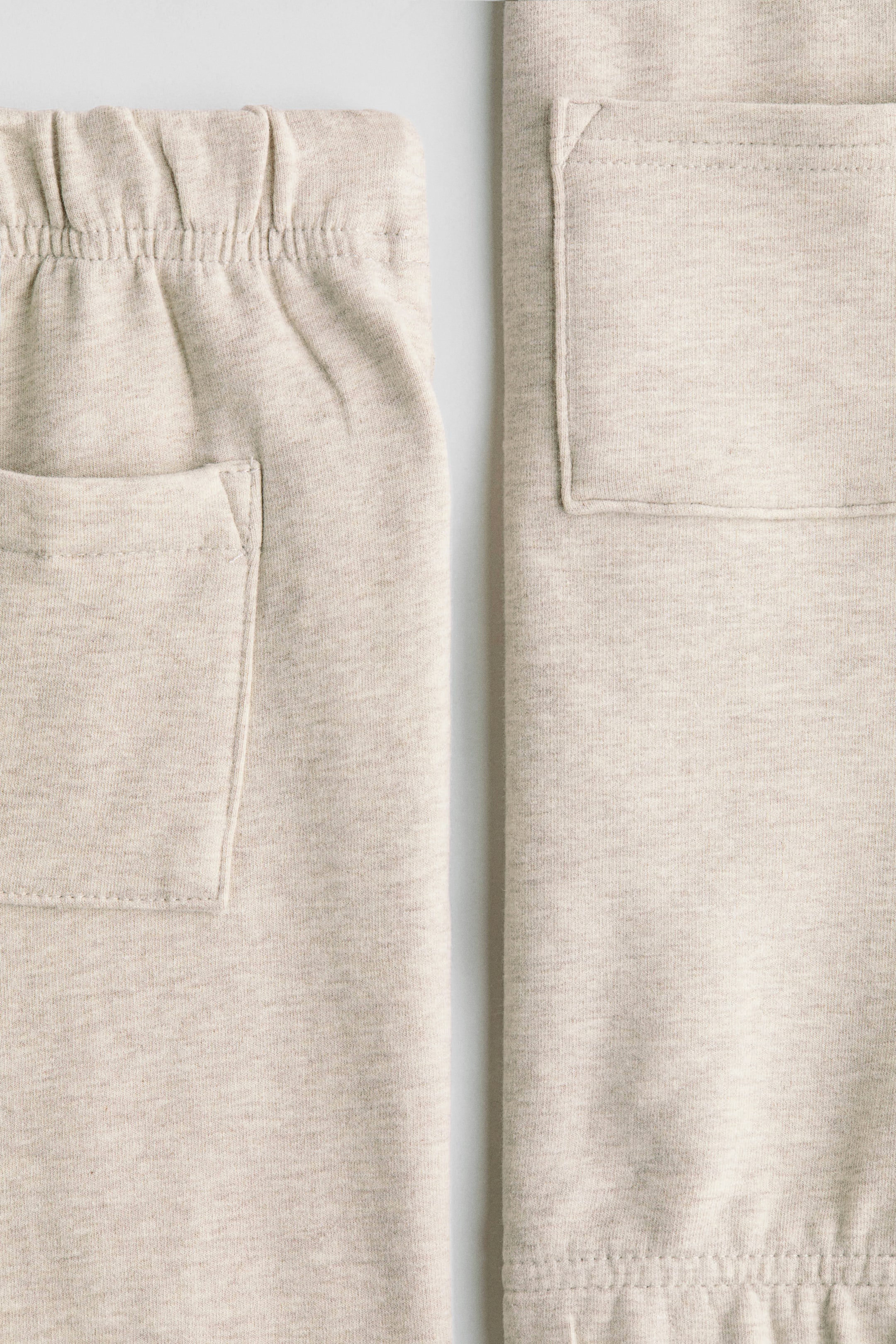 View larger image: A close-up of the H&M Adorables set is showing its light beige, heathered sweatshirt fabric. On the left, the front of the joggers is featuring an elasticated waistband with visible gathering and a rectangular patch pocket. On the right, a matching rectangular patch pocket and an elasticated hem are being displayed.